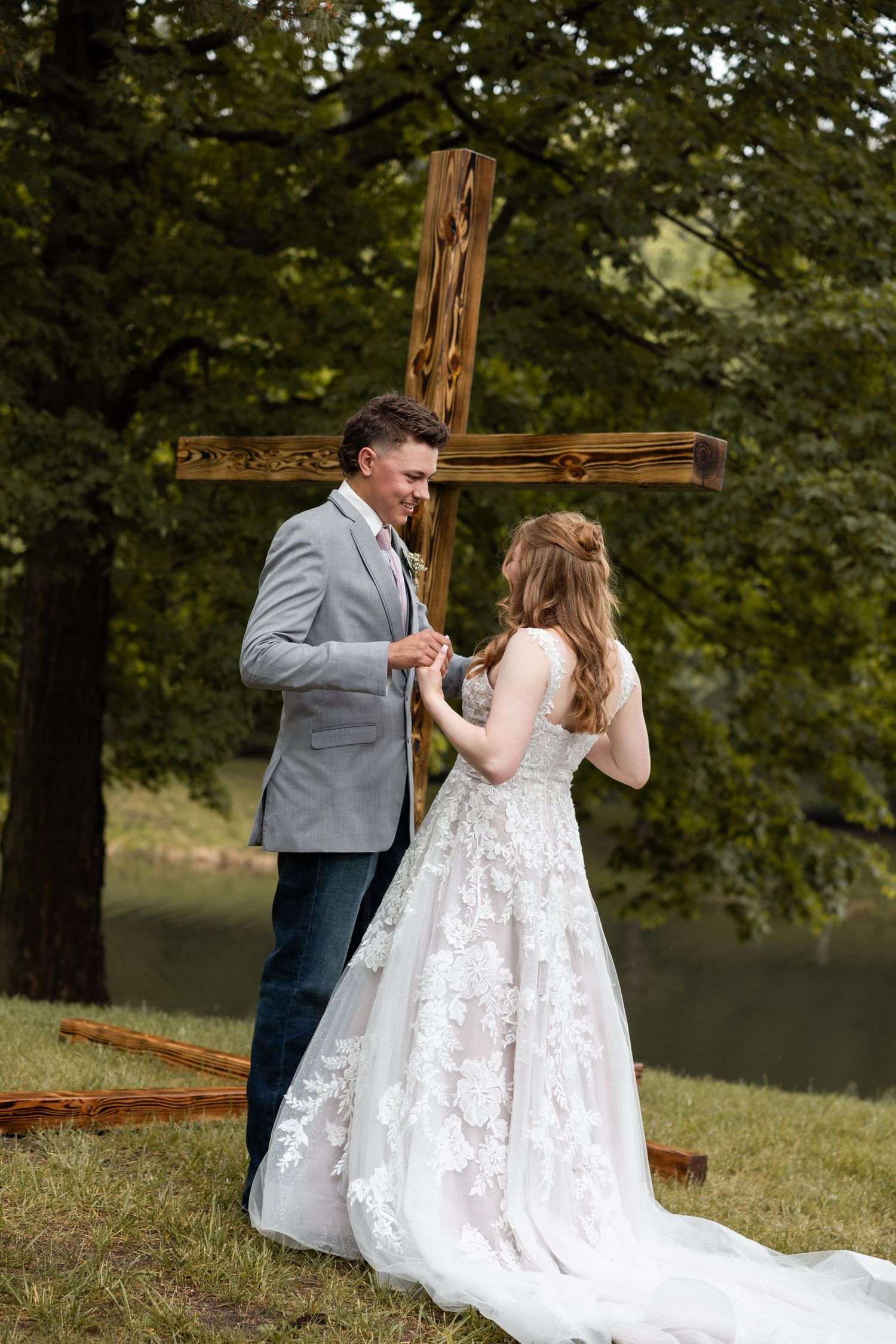Bride and groom exchange rings beside a large wooden cross in a lush outdoor setting.