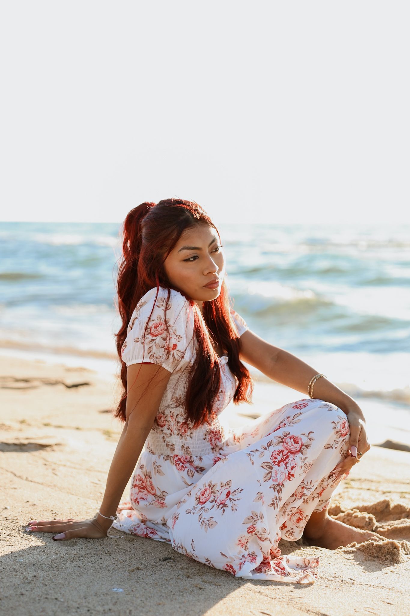 Young woman in floral dress sitting cross-legged on beach by ocean.