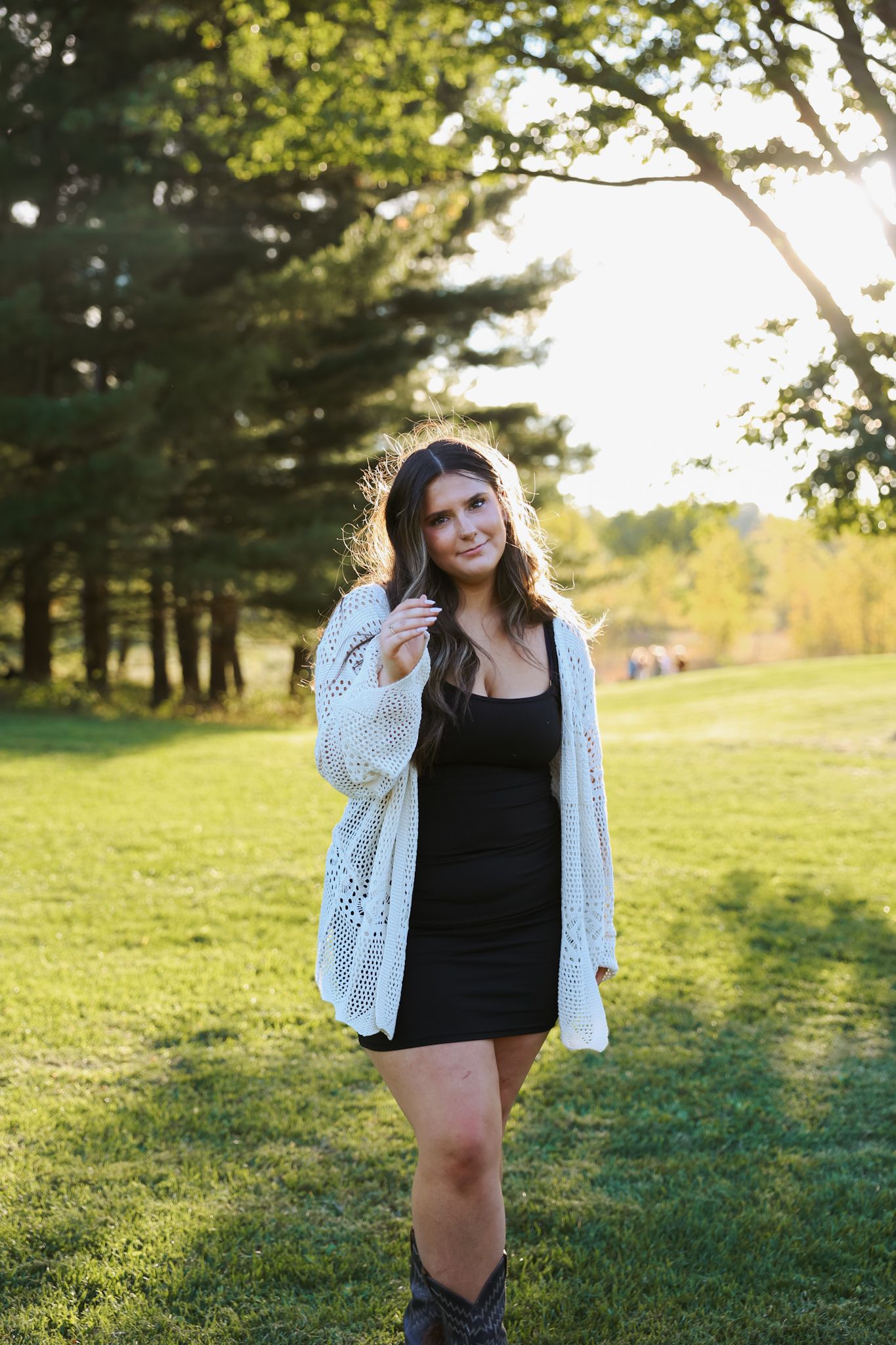 Young woman in white crochet cardigan and black mini dress standing on a sunny green lawn.