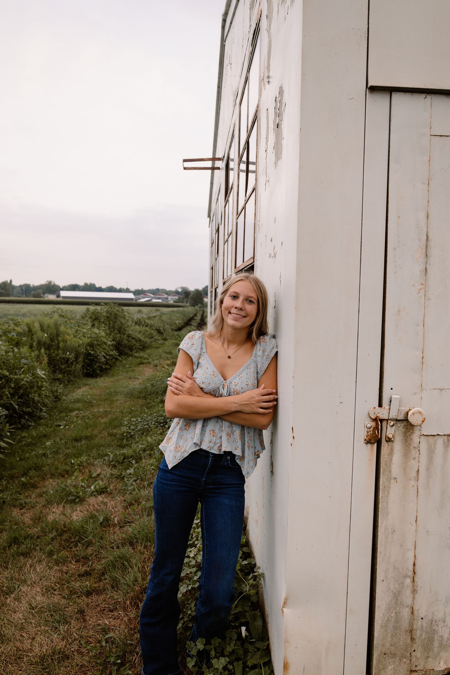 Young blonde woman in jeans and floral top leans against white barn wall in rural farm setting.