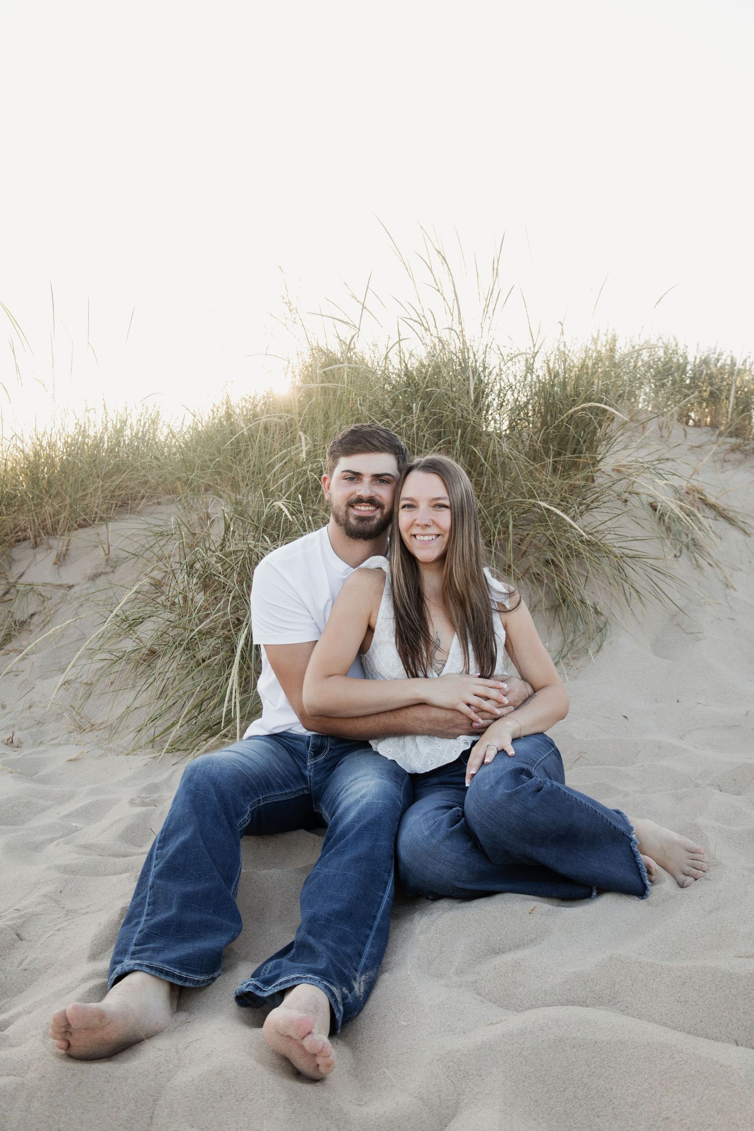 Couple sitting together on sandy beach dunes, smiling at camera, wearing white tops and jeans.
