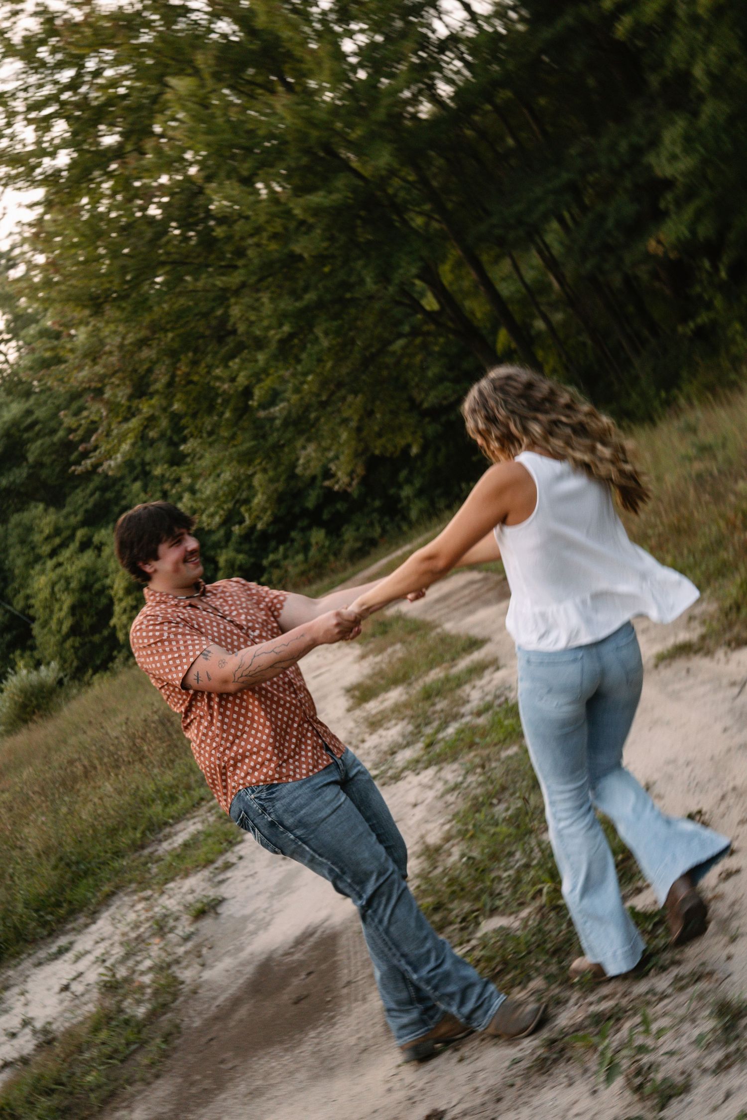 A couple dances and holds hands on a sandy path surrounded by lush green trees at golden hour.