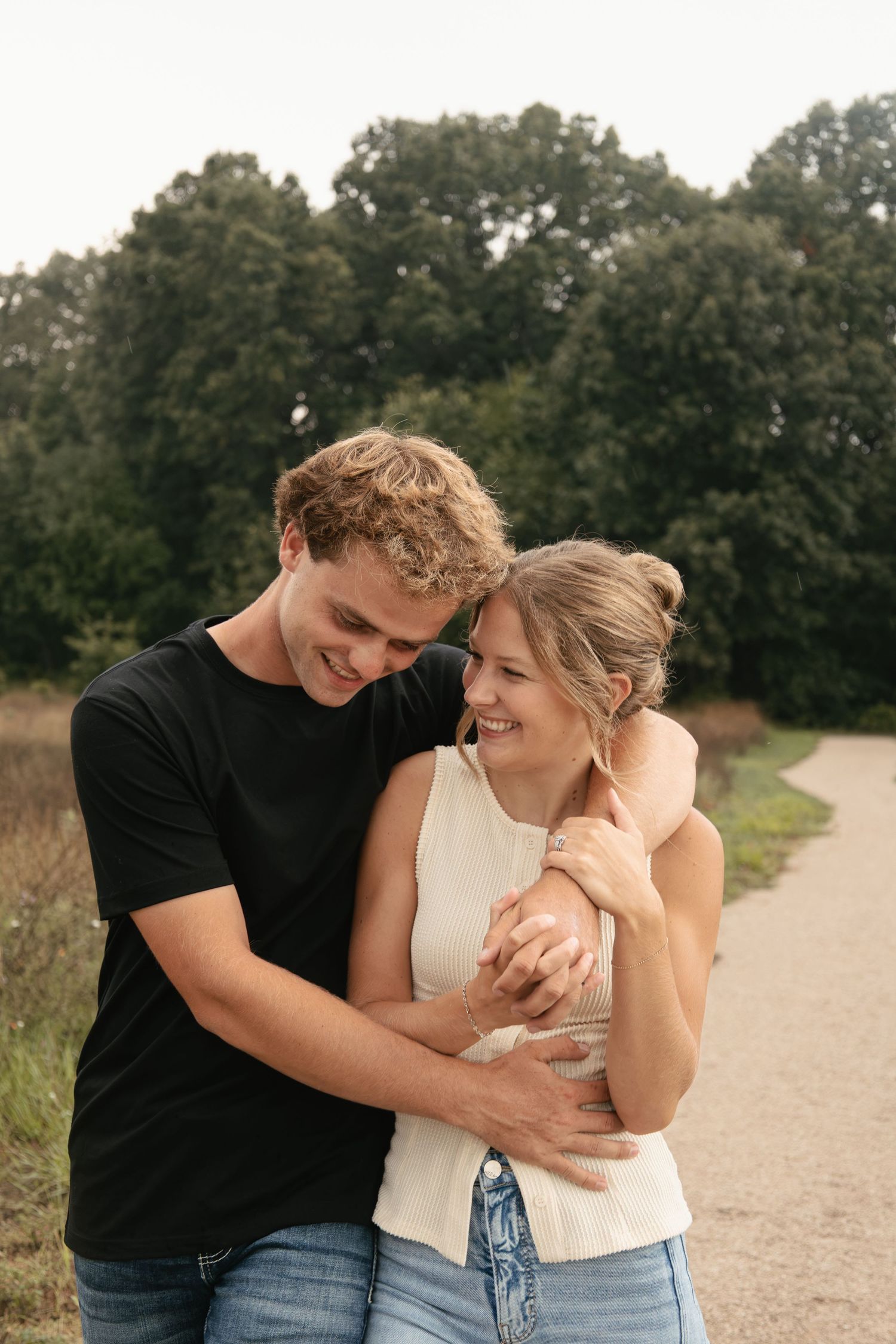 Happy couple embracing on a scenic path surrounded by lush green trees, man in black tee hugging woman in white top.