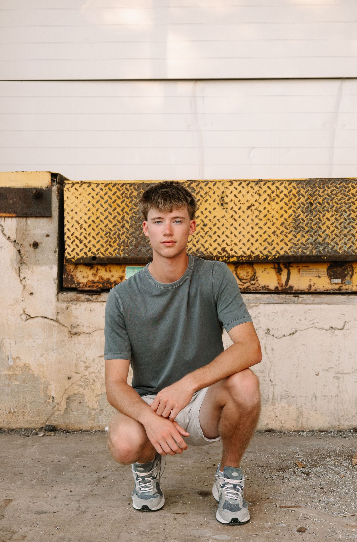 Young man in gray t-shirt and shorts crouching in front of a rustic industrial wall with yellow diamond plate metal.