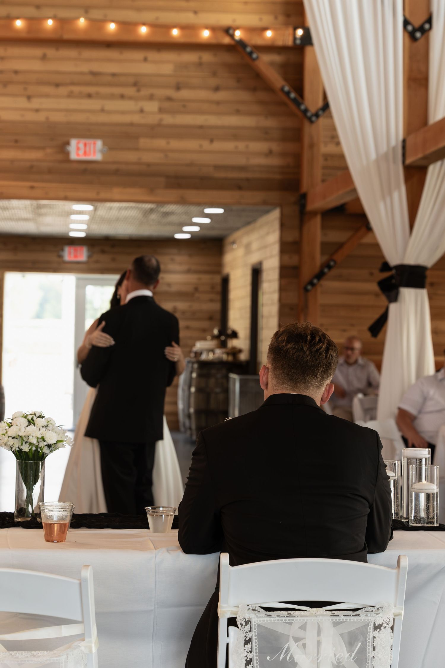 Groom sits at head table watching couple dance in elegant rustic barn wedding reception venue.