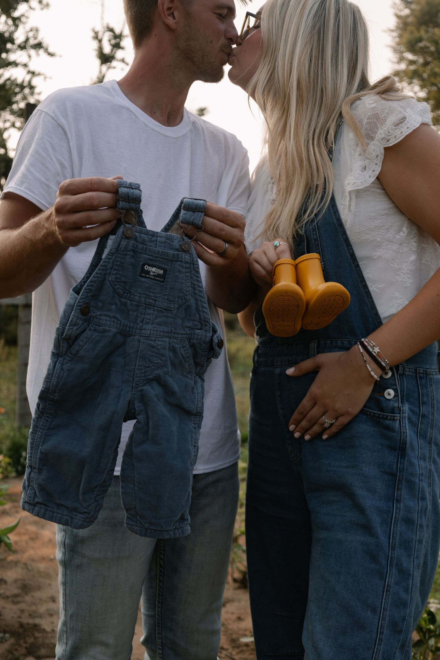 Couple kissing while holding tiny denim overalls and yellow boots, announcing pregnancy outdoors at golden hour.