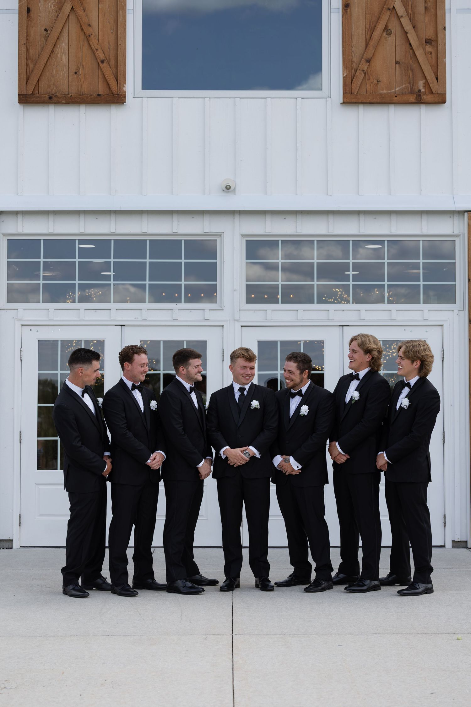 Seven groomsmen in black tuxedos with white boutonnieres stand in a semi-circle outside a white barn venue.