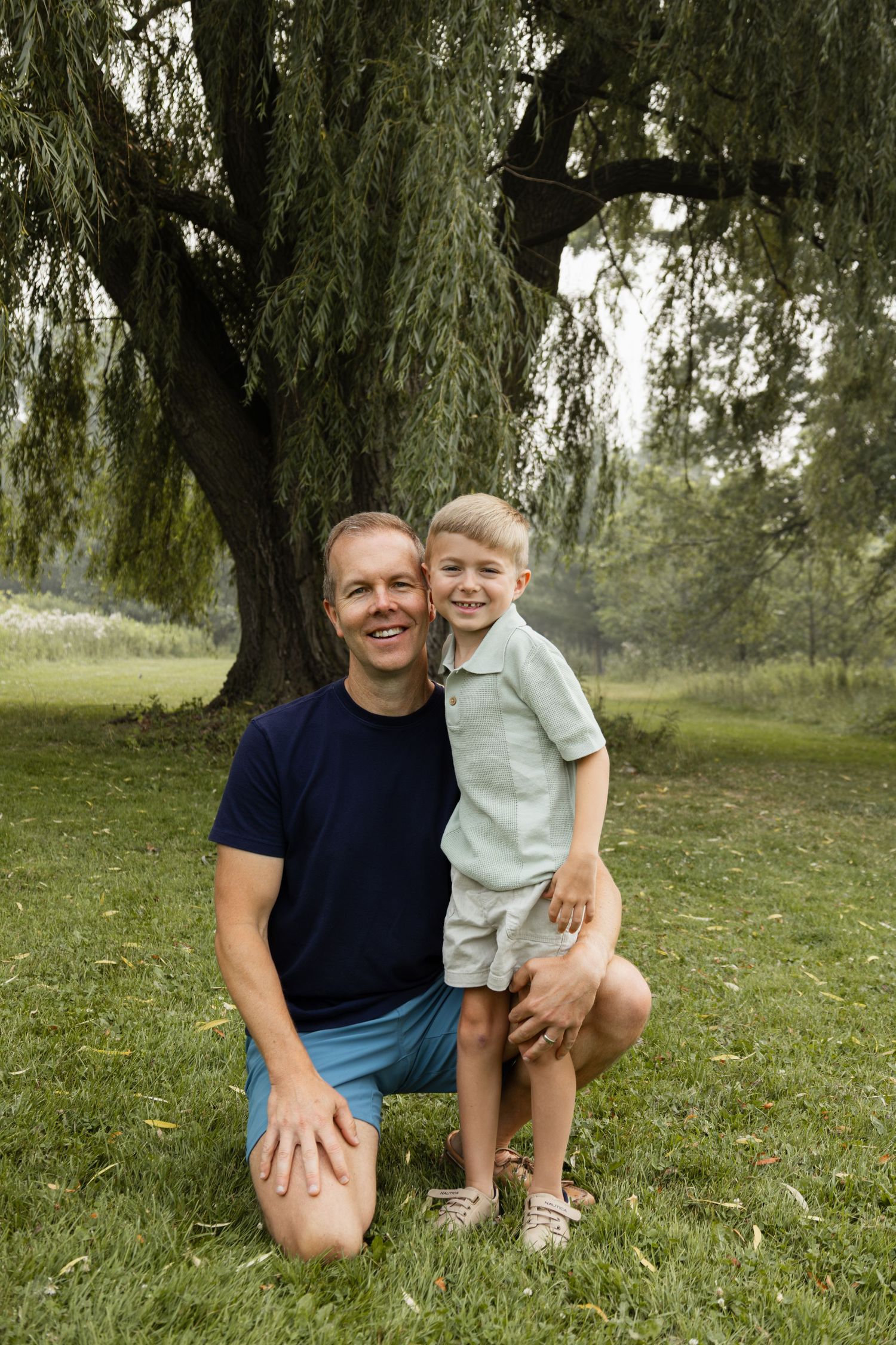 A smiling man kneeling beside a young boy under a large willow tree in a lush green park setting.
