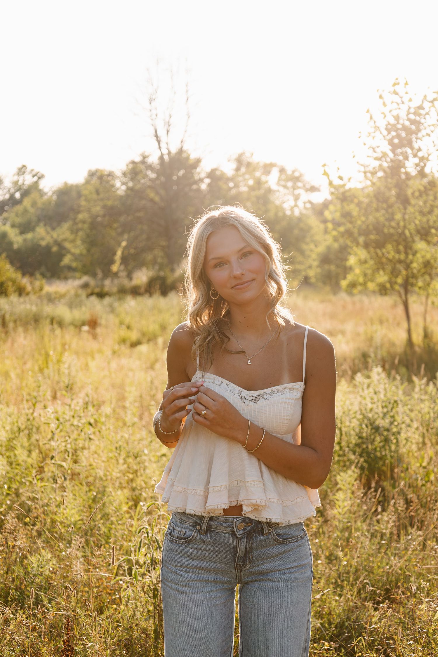 Young blonde woman in white lace crop top and jeans standing in sunlit meadow with golden hour backlight.