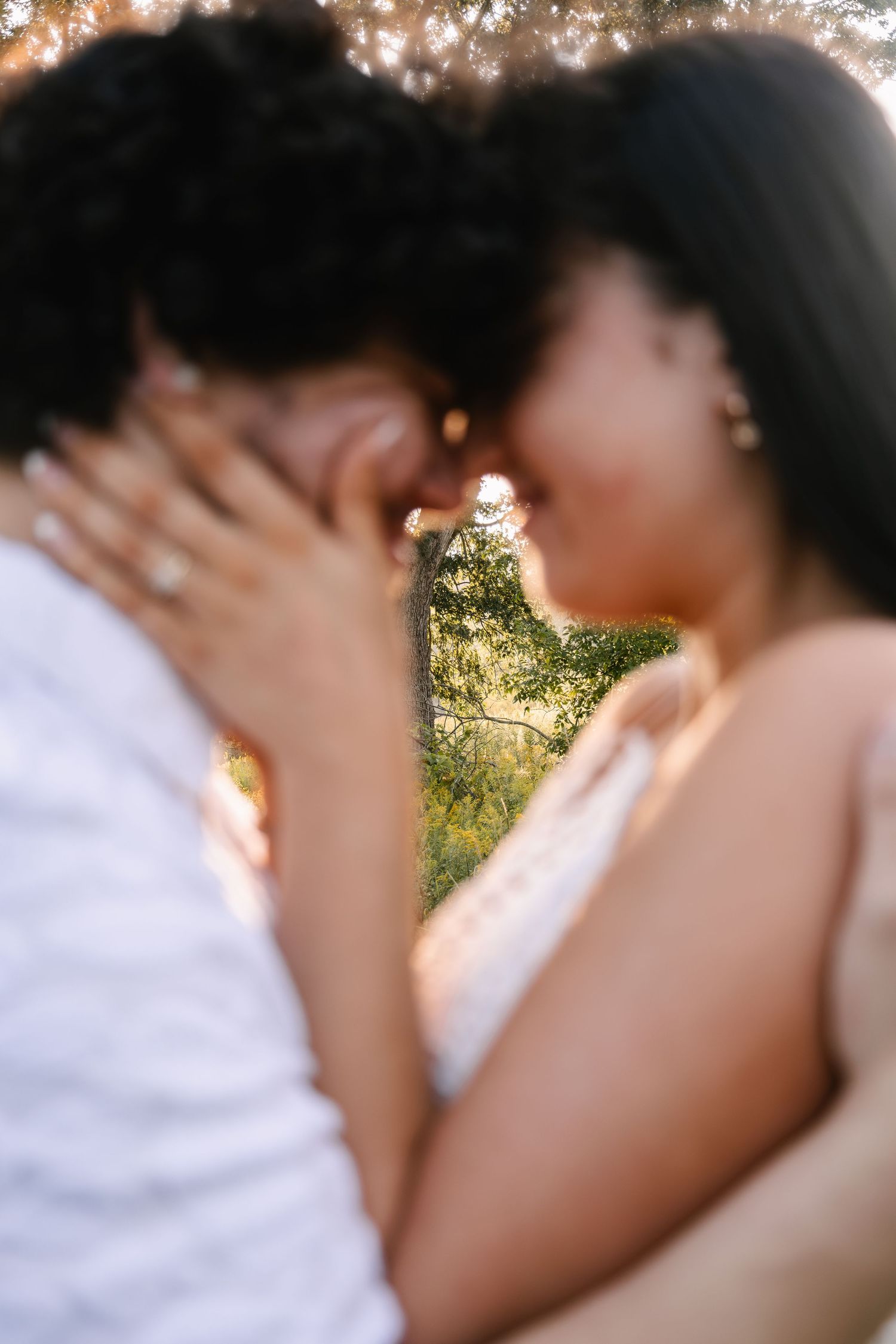 Couple sharing a romantic kiss outdoors in golden sunlight, with lush green trees in the background.