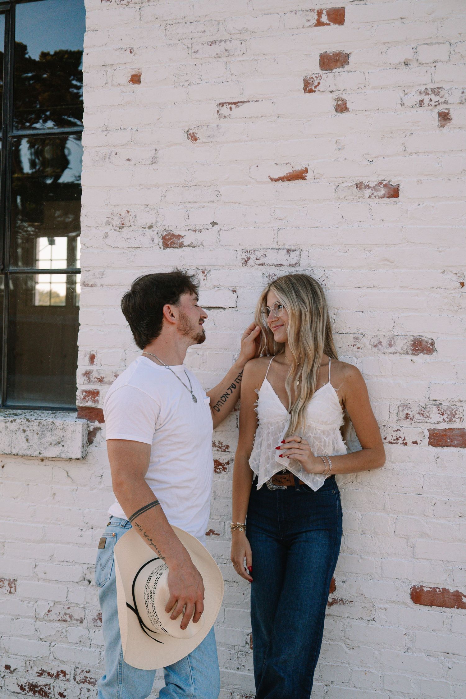 A couple smiles at each other against a white brick wall, man holding a cowboy hat in jeans and white tee.
