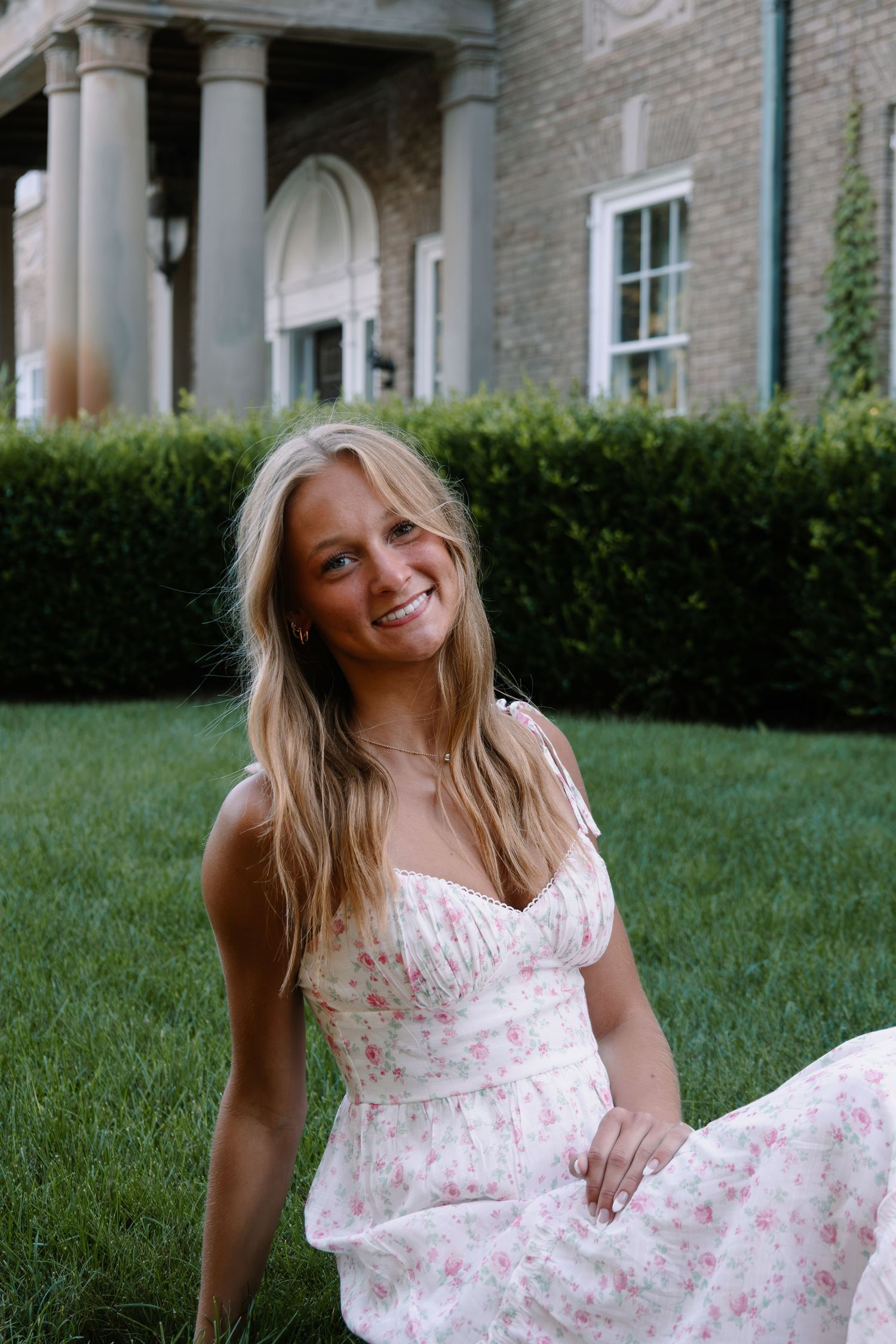 Smiling blonde woman in floral sundress sitting on green grass in front of elegant brick building with columns.
