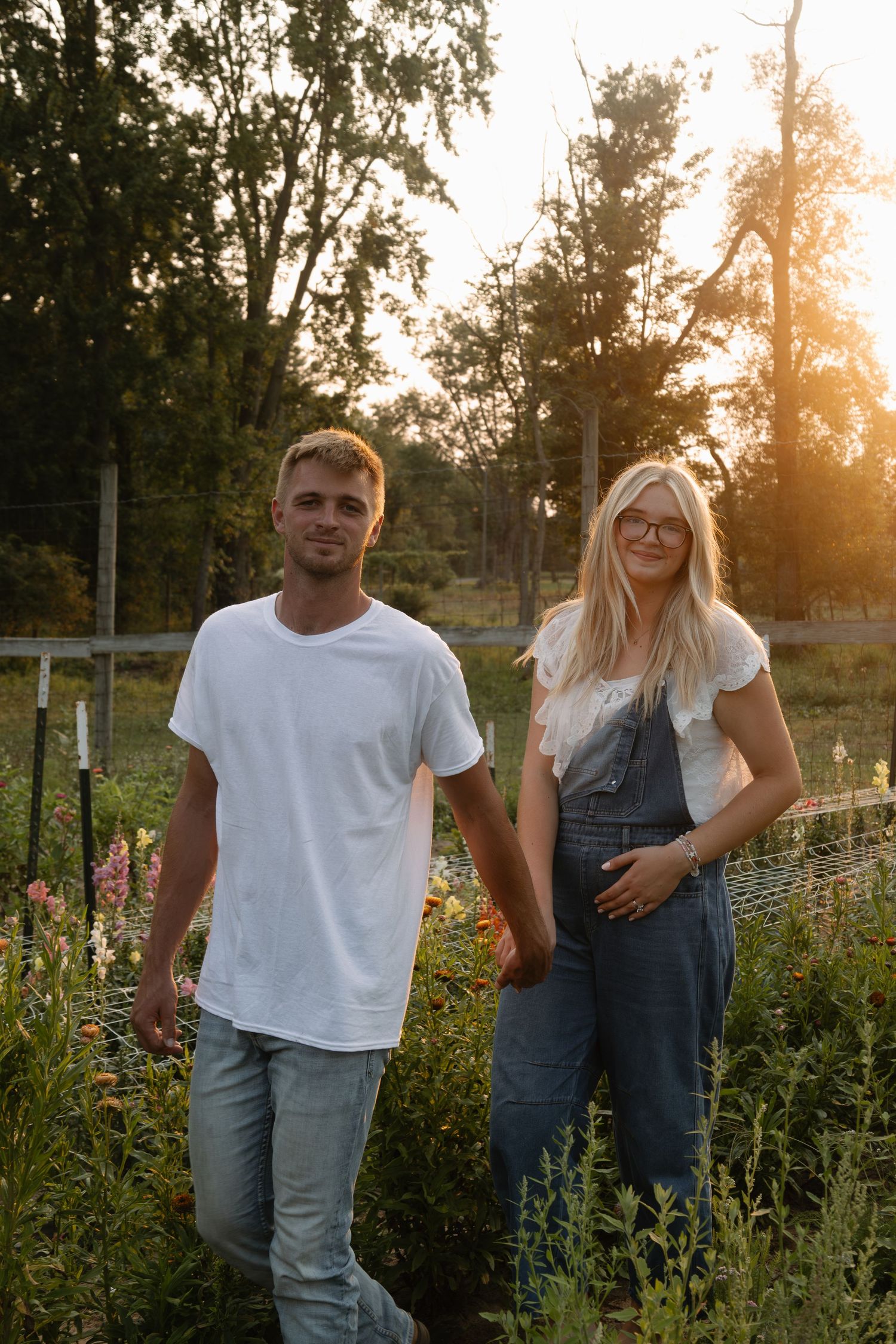 A young couple holds hands in a sunlit garden at golden hour, surrounded by wildflowers and lush greenery.