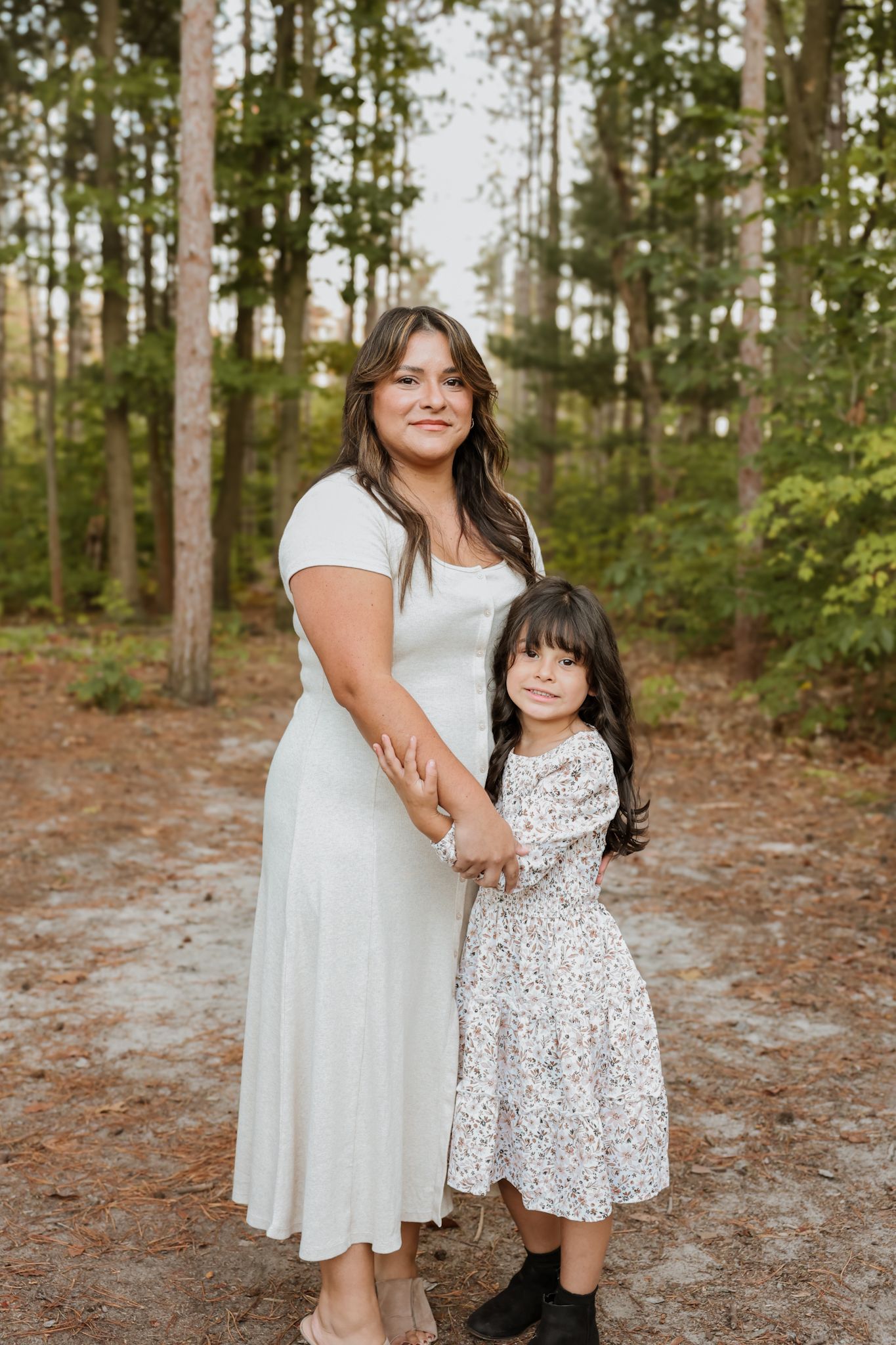 Mother in white dress poses with young daughter in floral dress in a pine forest.