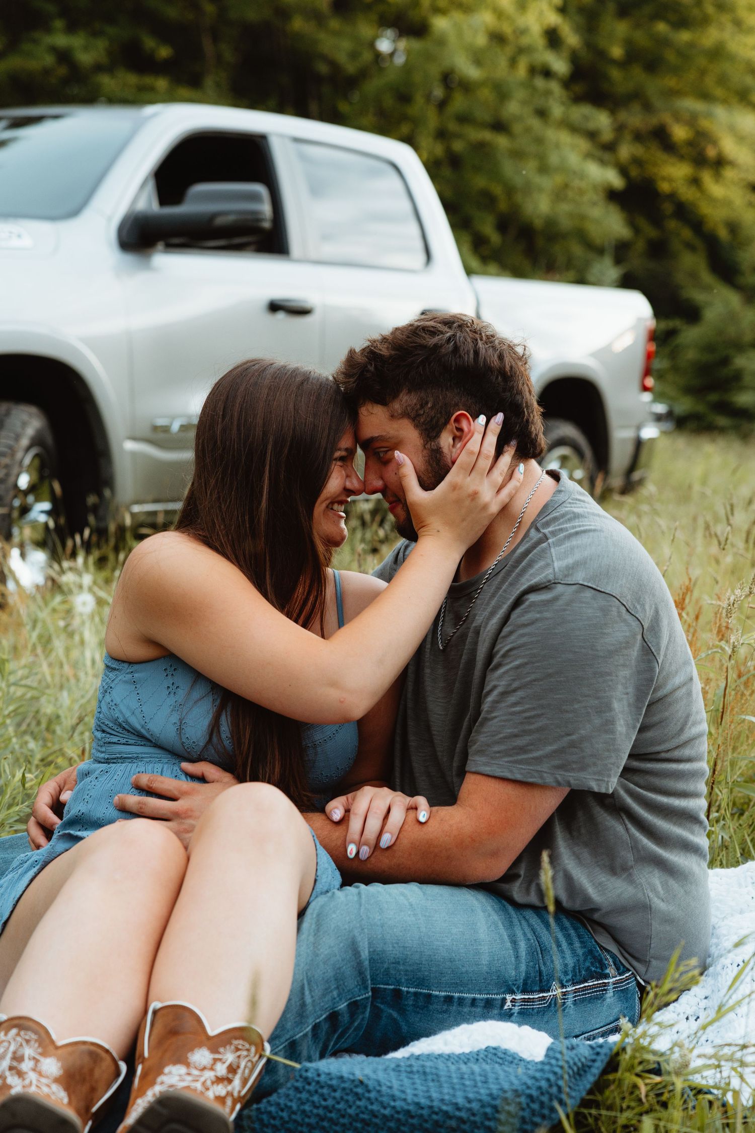 Couple sitting together in tall grass, sharing a tender forehead-to-forehead moment by a truck.