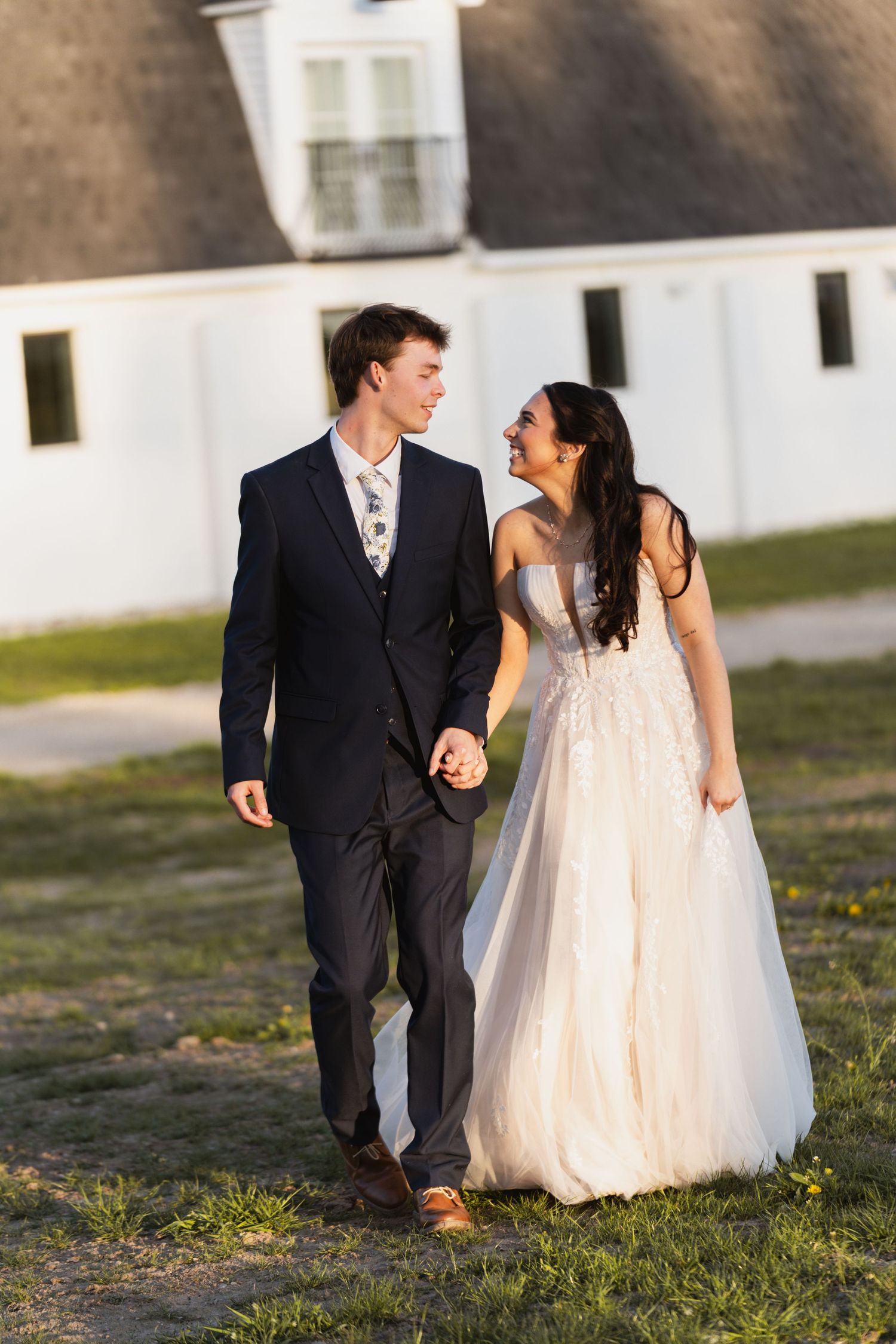 Happy bride and groom walk hand in hand across a green lawn with a white barn in the background.
