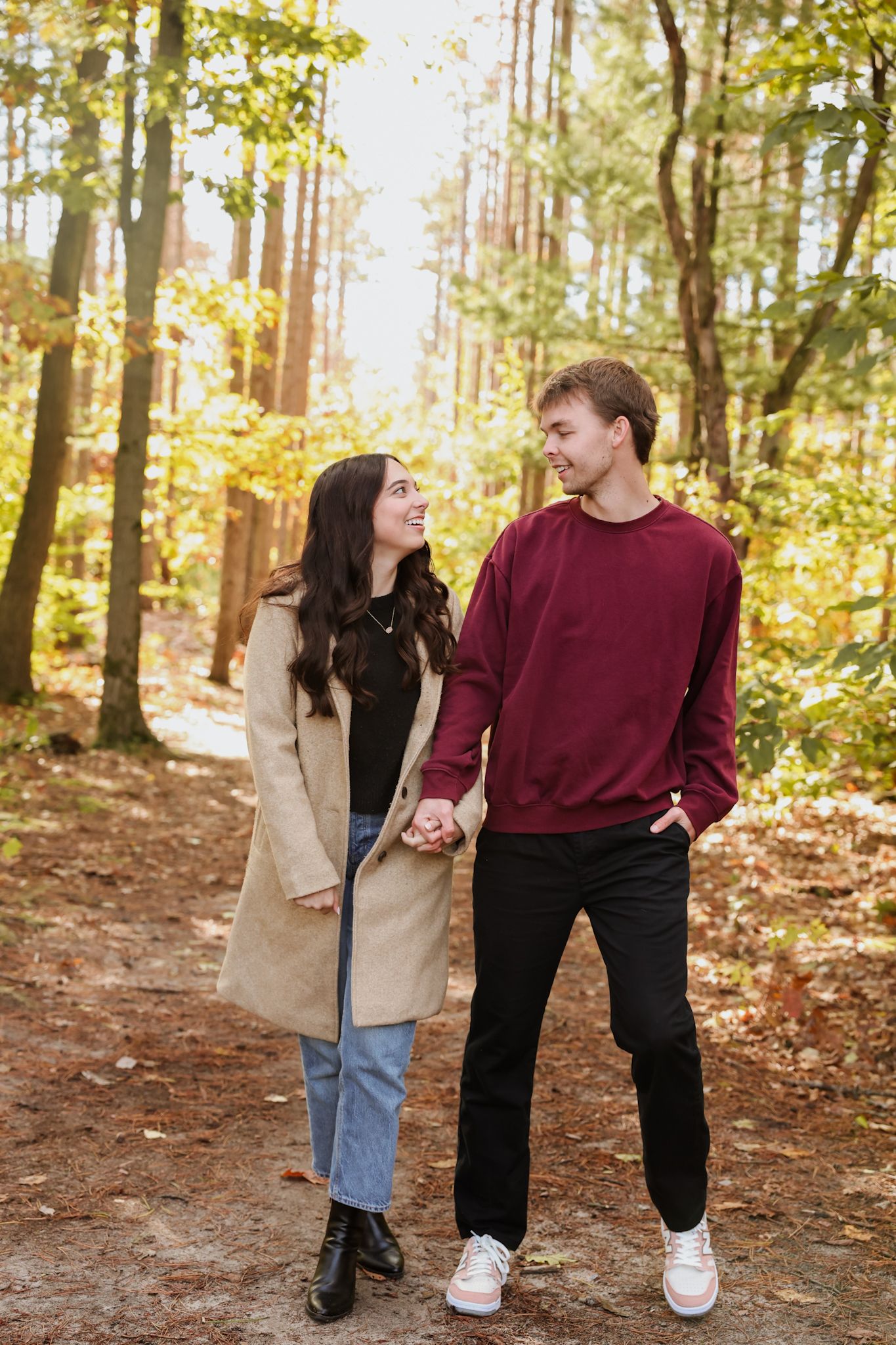 A happy couple holds hands while walking on a forest path surrounded by colorful autumn foliage.
