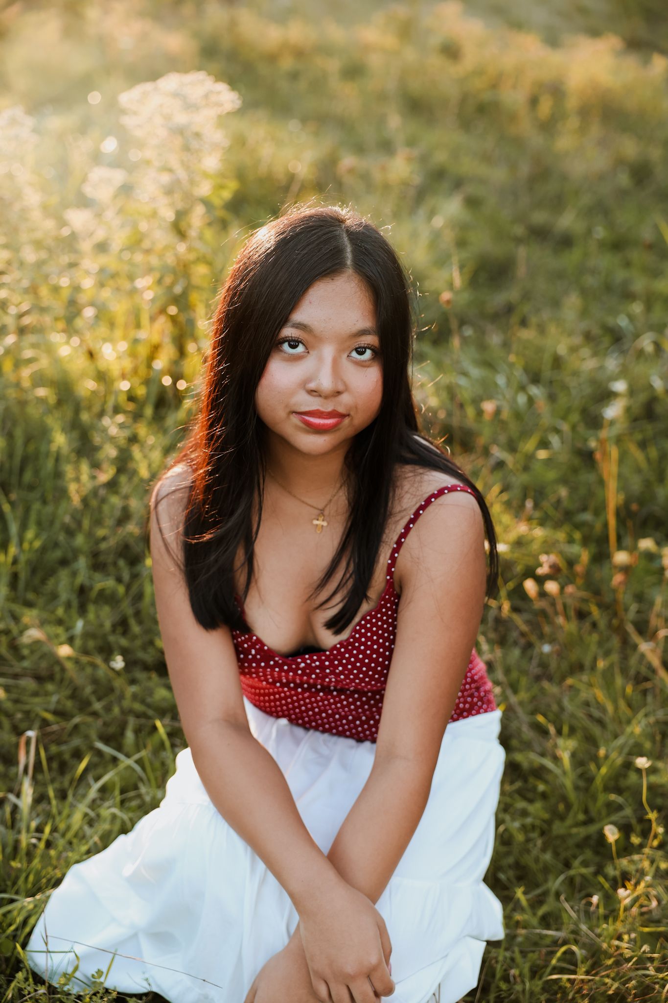 Young Asian woman in red polka dot top sits in sunlit wildflower meadow for senior portraits.