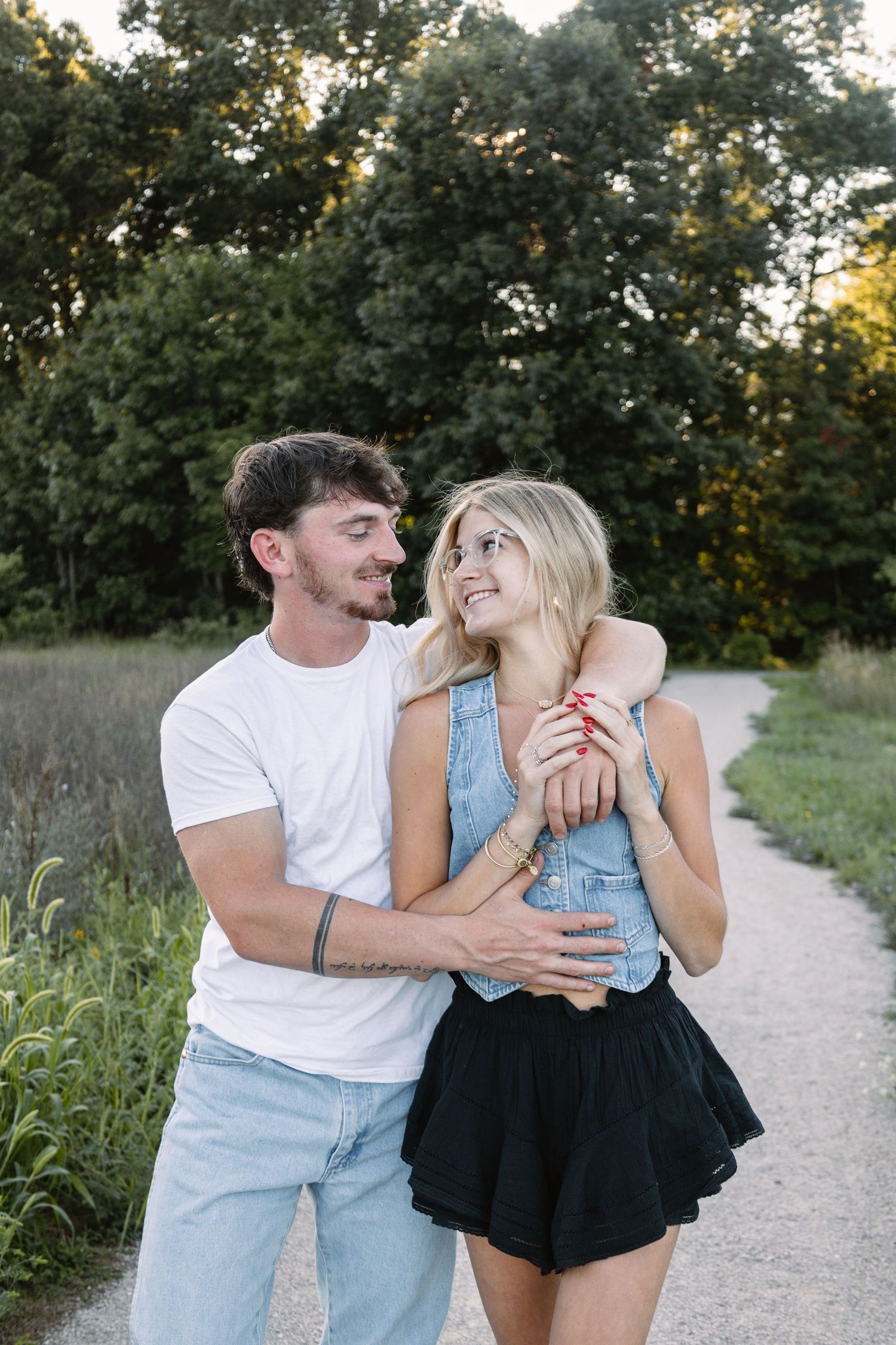 A couple embraces on a scenic path surrounded by lush green trees, smiling at each other in a romantic outdoor setting.