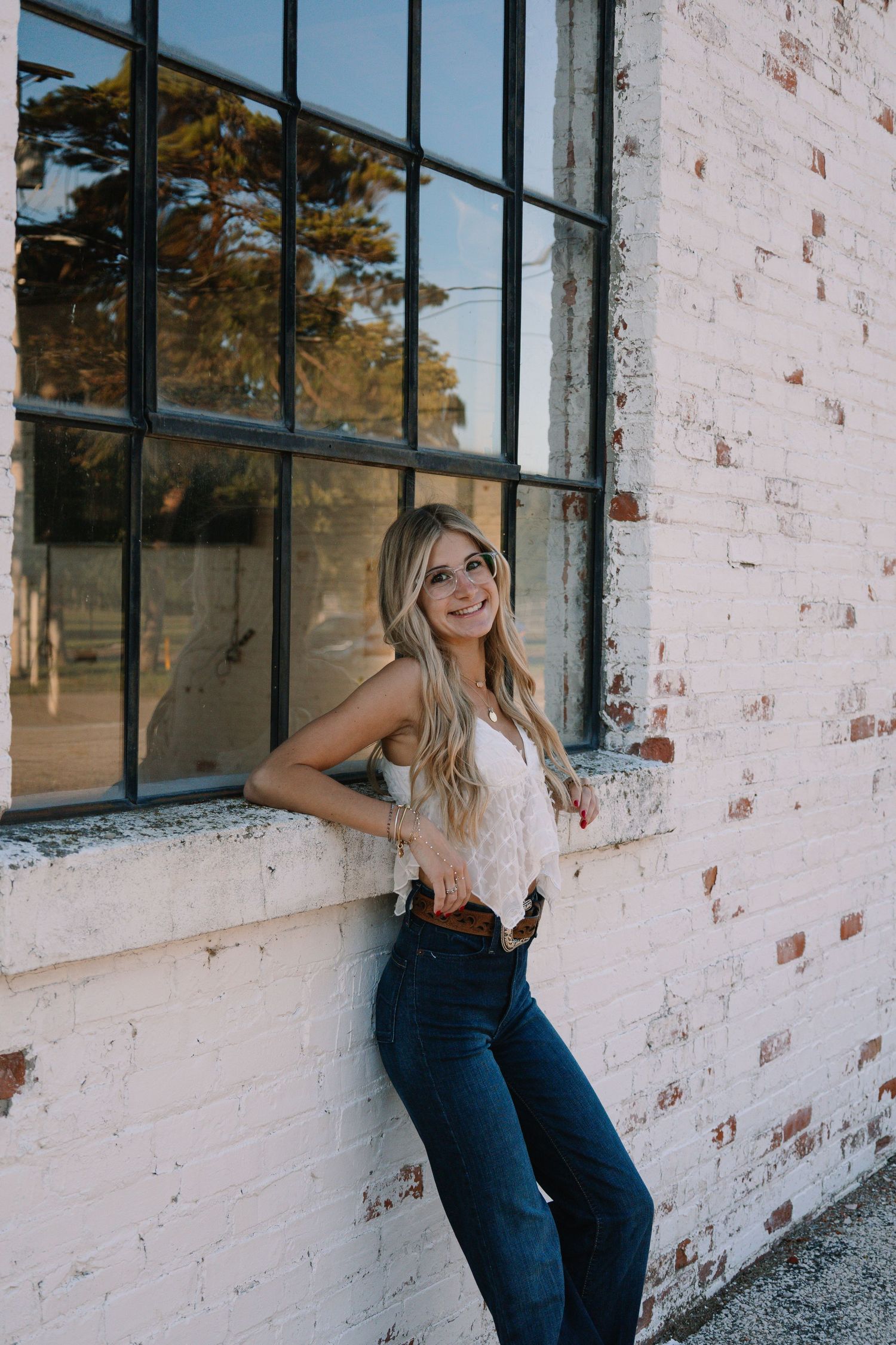 Smiling blonde woman in jeans and white top leaning against a white brick wall by a large window.