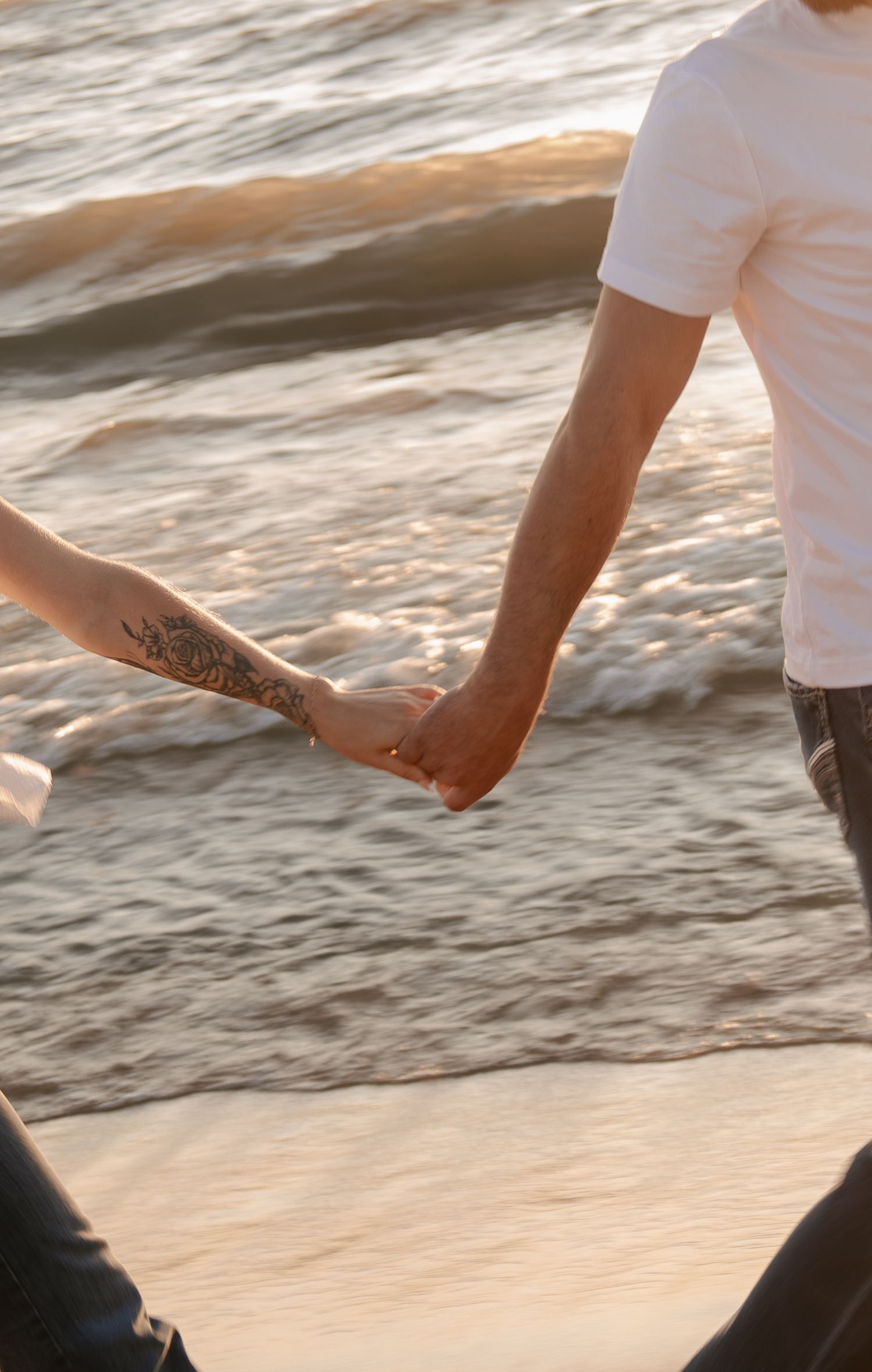 Couple holding hands on a sandy beach at sunset, one partner showing a floral forearm tattoo.