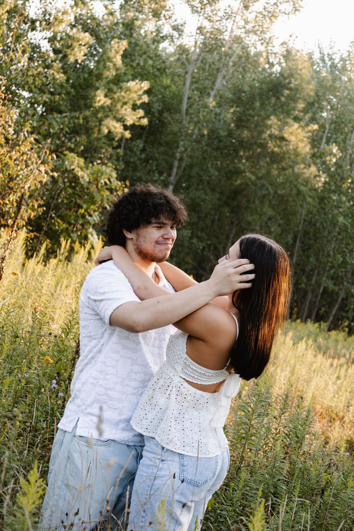 A happy couple holds each other closely in a sunlit meadow surrounded by lush green trees.