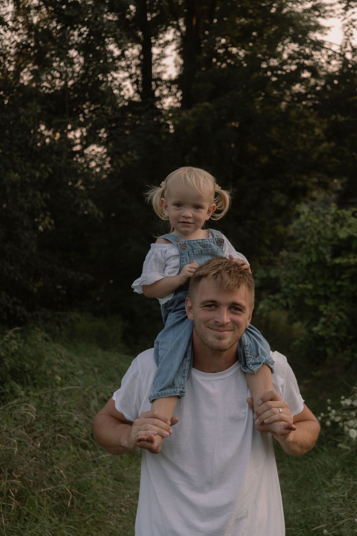 A smiling young man in a white t-shirt carries a happy toddler girl on his shoulders outdoors.