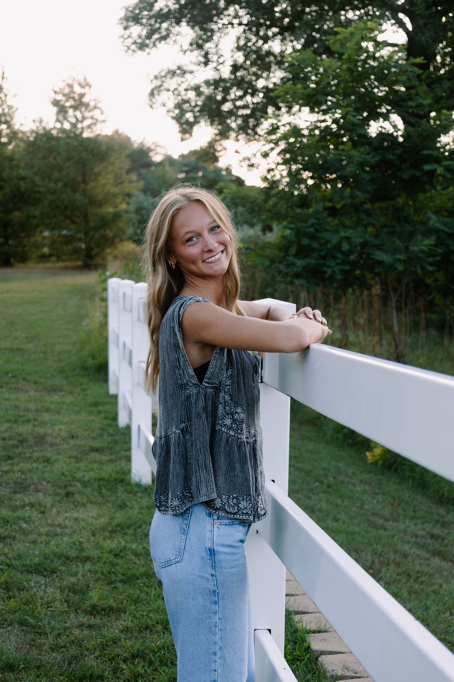 Smiling blonde woman leaning on white fence in green field wearing denim vest and jeans during golden hour.