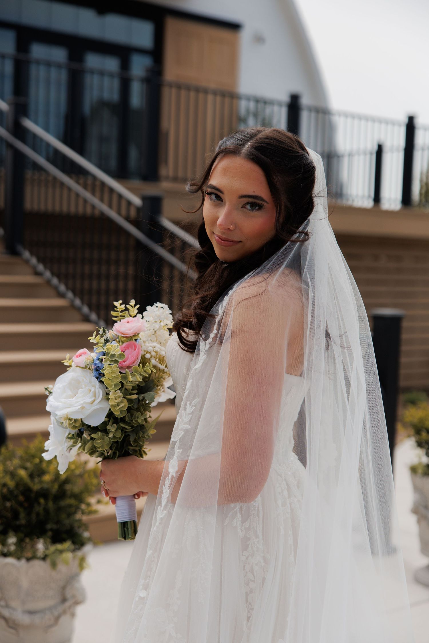 Elegant bride in white lace gown and veil holding a pink and white floral bouquet outdoors.