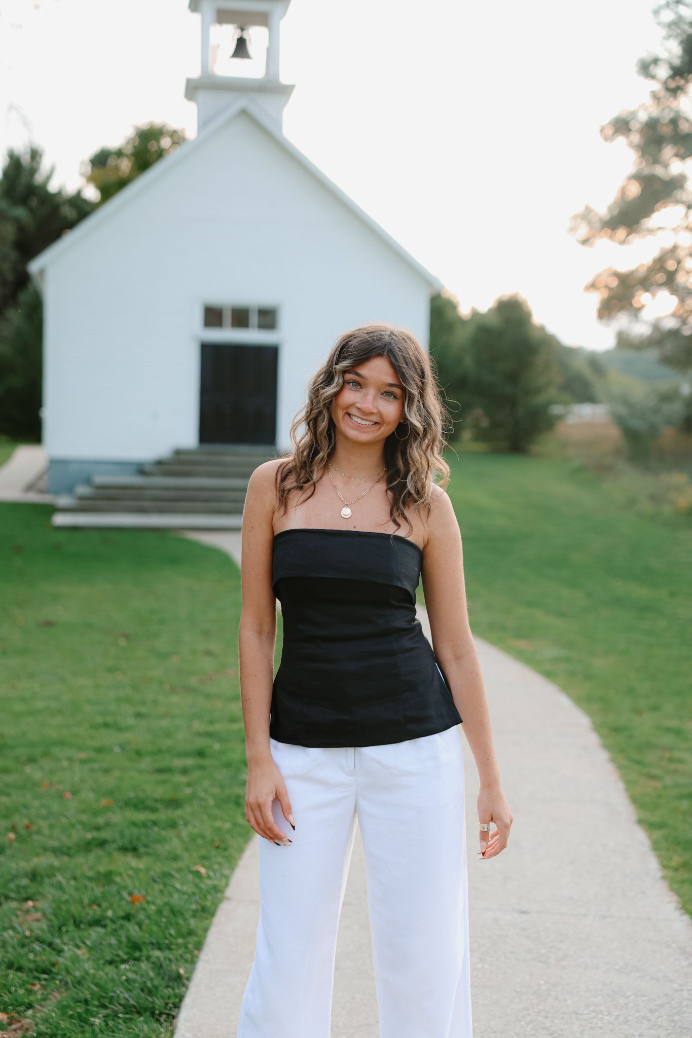Smiling young woman in black tube top and white pants standing on pathway in front of a white chapel.