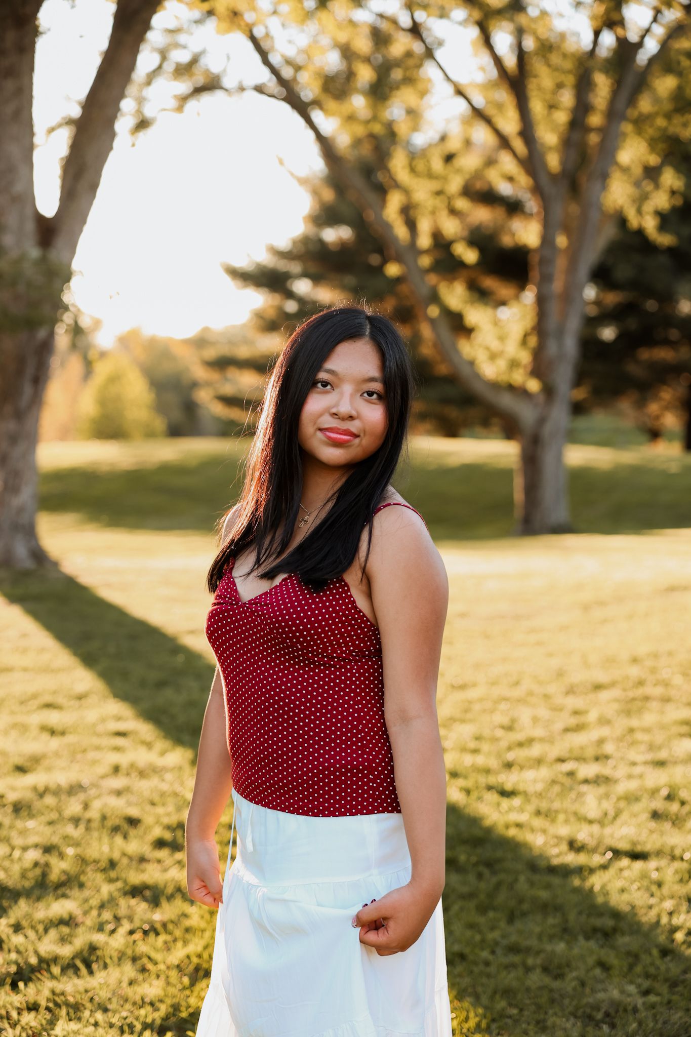 Young Asian woman in red polka dot cami and white skirt smiles in a sunny park at golden hour.