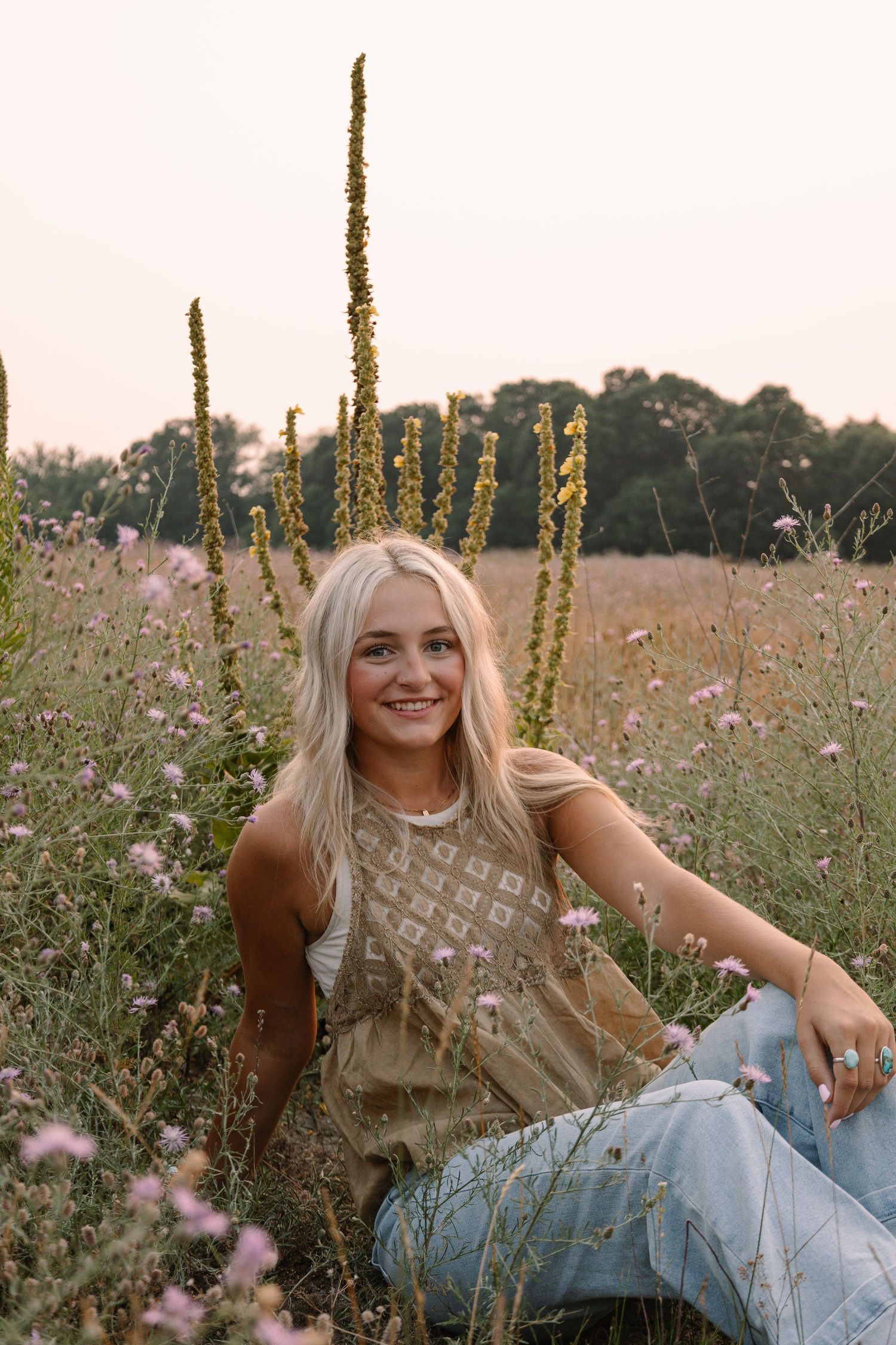 Smiling blonde woman sitting in a wildflower meadow wearing a boho top and jeans at golden hour.