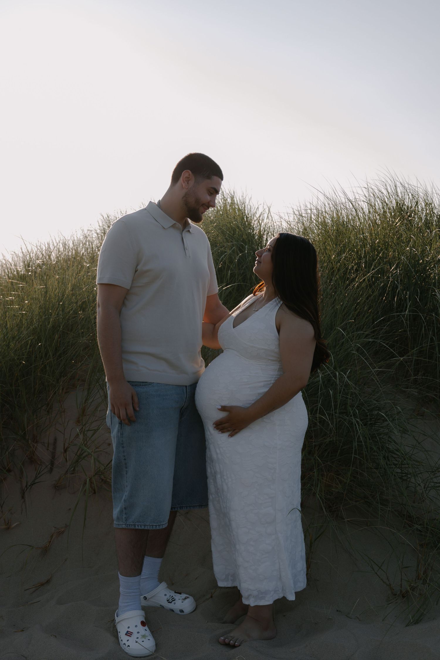 Pregnant couple sharing a tender moment on beach dunes at sunset, woman in white dress cradling baby bump.
