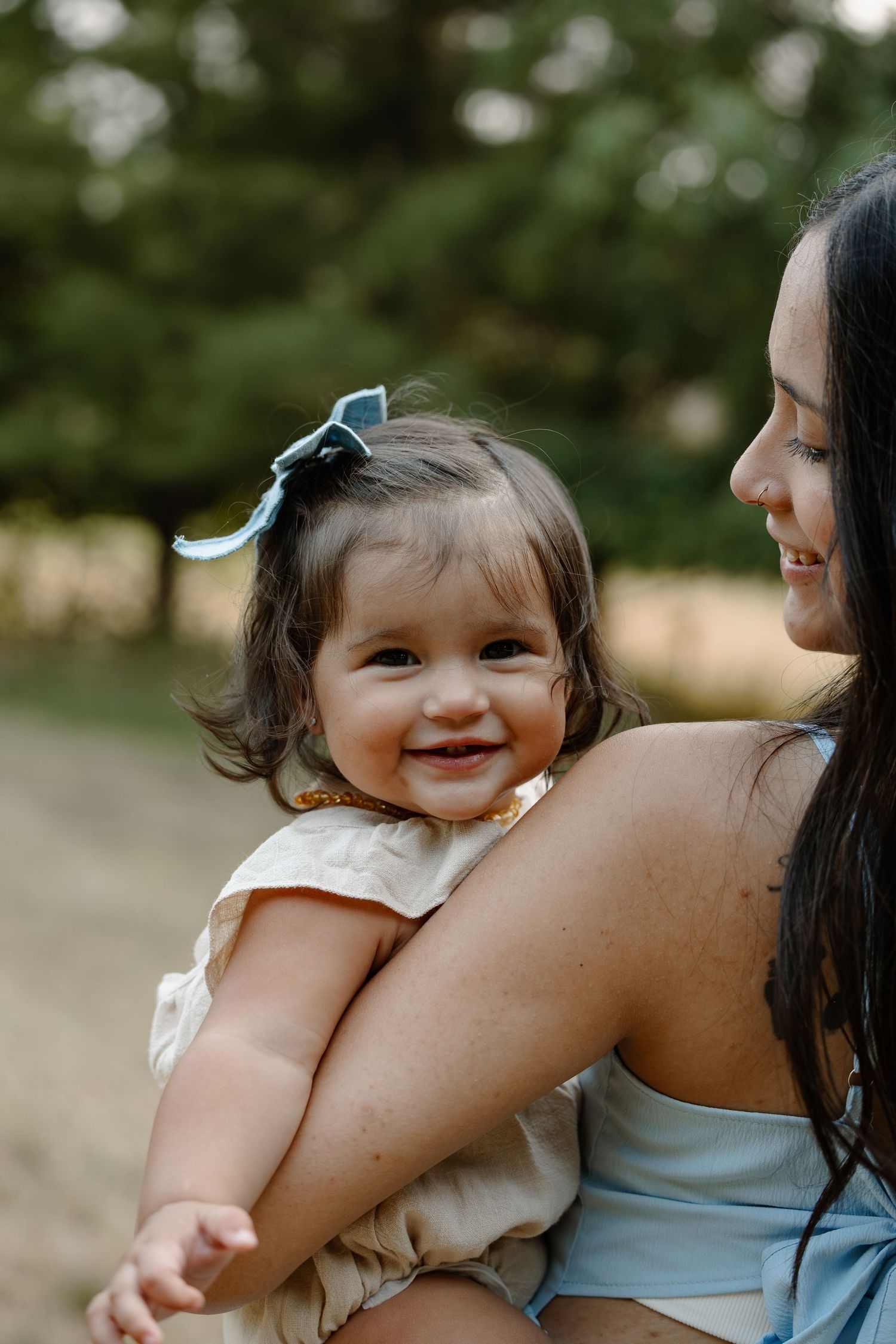 A smiling baby girl with a blue bow sits in her mother's arms outdoors near green trees.