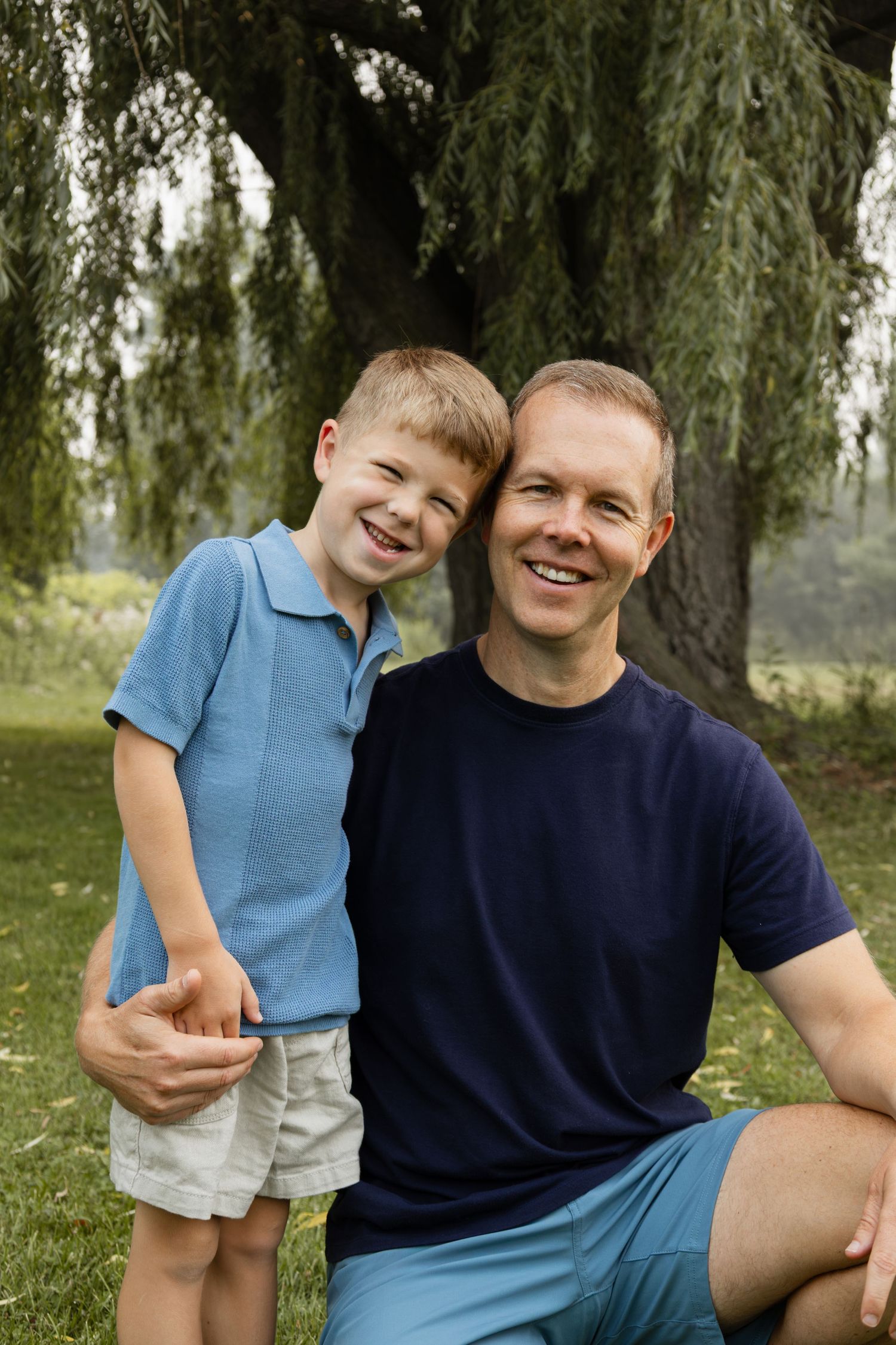 A smiling father and young son pose together outdoors near a willow tree on a sunny day.