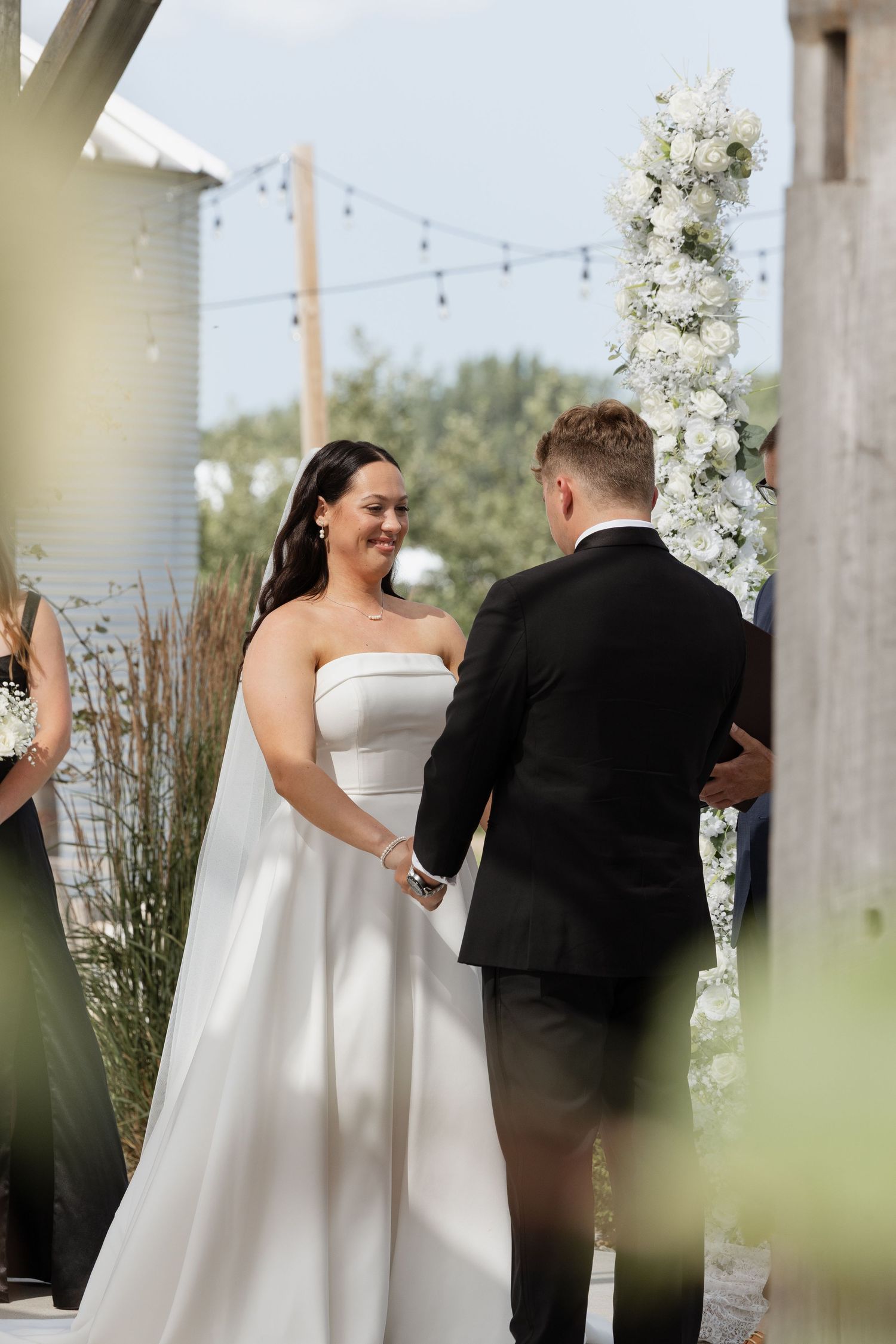 Smiling bride in strapless white gown holds hands with groom at outdoor wedding ceremony near floral arch.