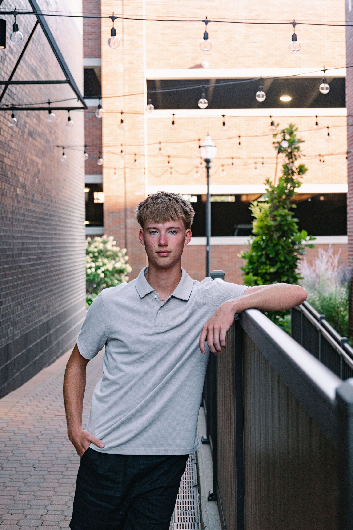 Young man in gray polo leans on railing in urban courtyard with string lights and brick walls.