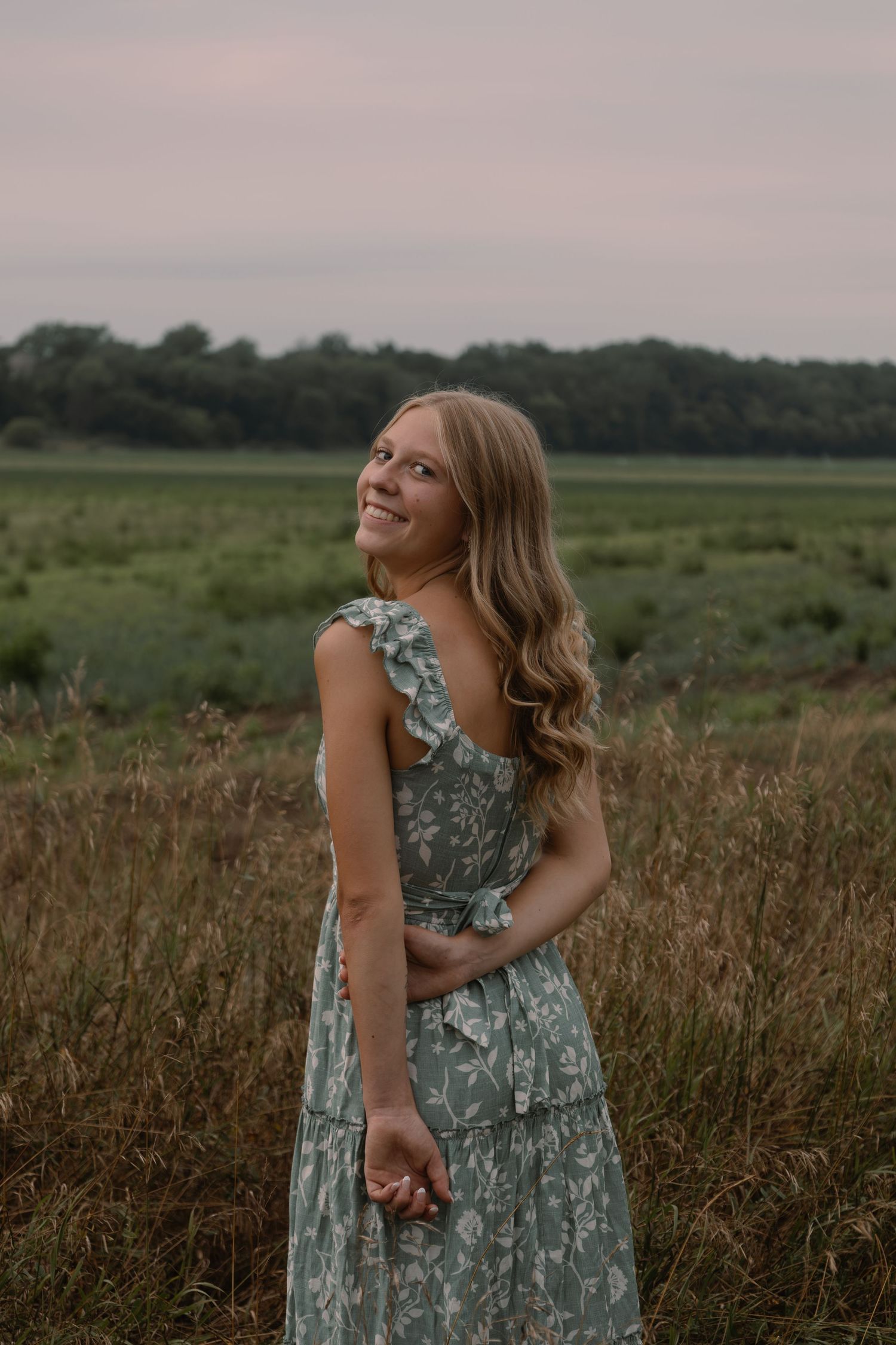 Young woman in floral sage green dress smiling over shoulder in golden meadow at dusk.