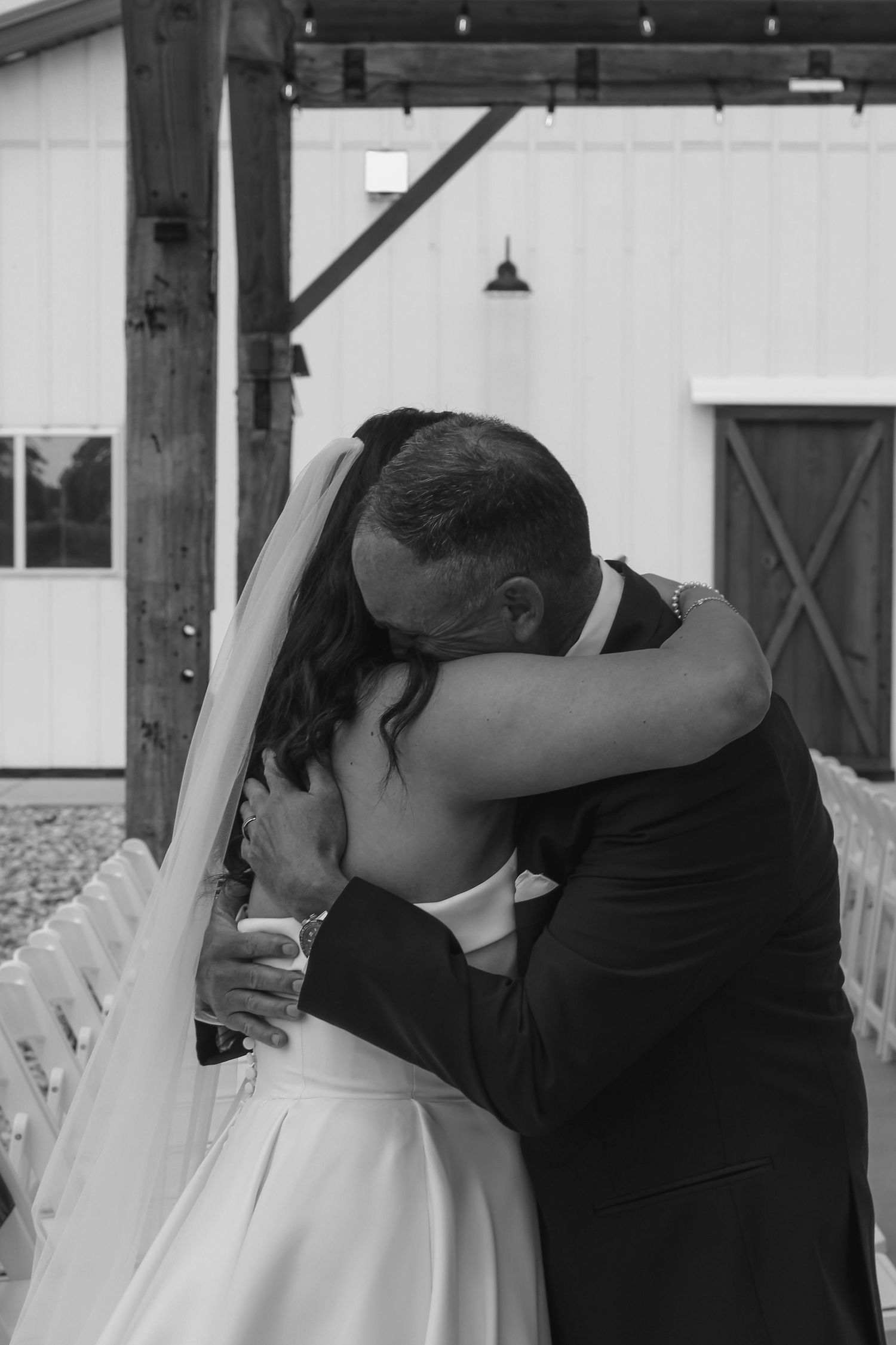 Black and white photo of a bride and groom sharing an emotional first kiss embrace at an outdoor barn wedding ceremony.