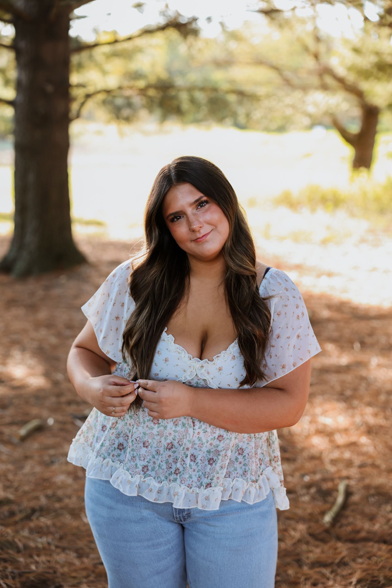 Young woman in floral lace top and jeans posing outdoors near trees in golden hour light.