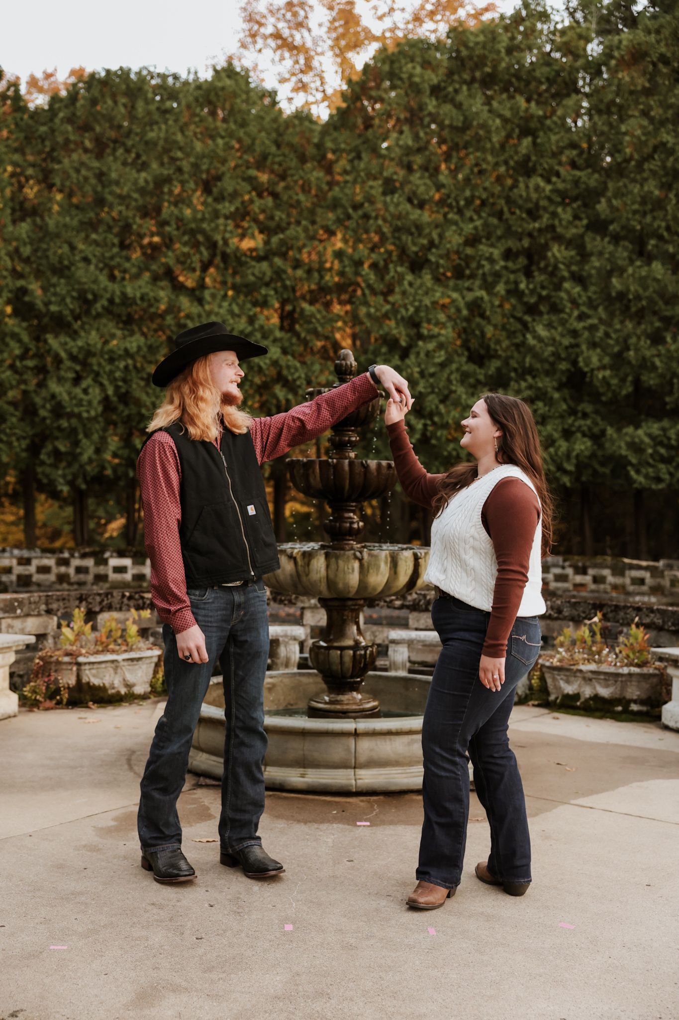 Fun couple dancing and spinning near a stone fountain surrounded by colorful autumn foliage.