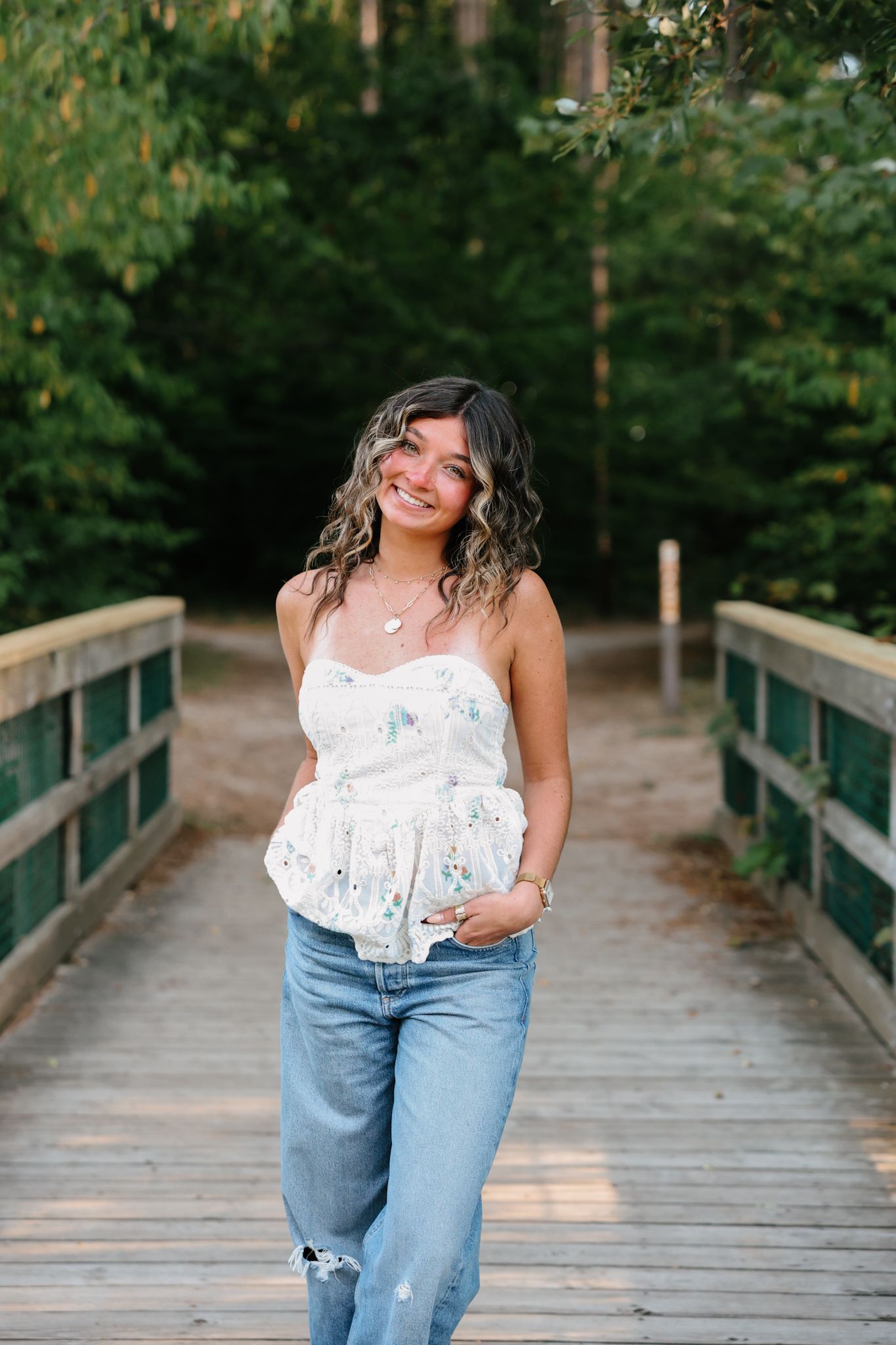 Smiling young woman in floral top and jeans walking on wooden bridge.