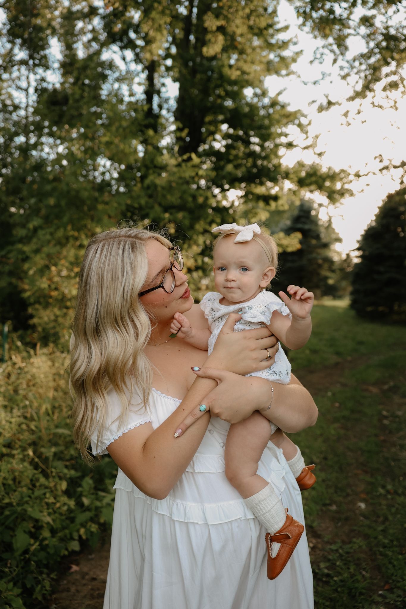 Blonde mother in white dress holding baby girl in floral romper in beautiful outdoor garden setting.