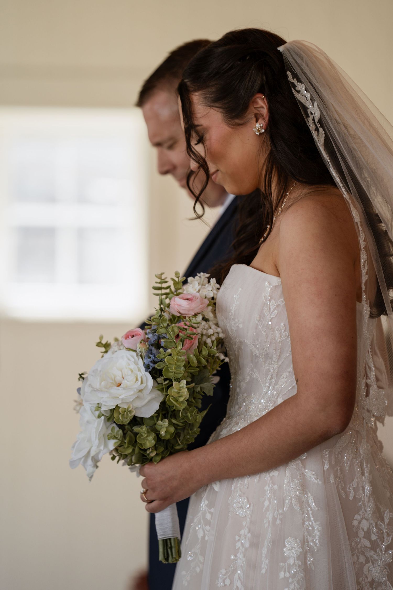 Emotional bride in lace gown holds pink and white bouquet during indoor wedding ceremony.