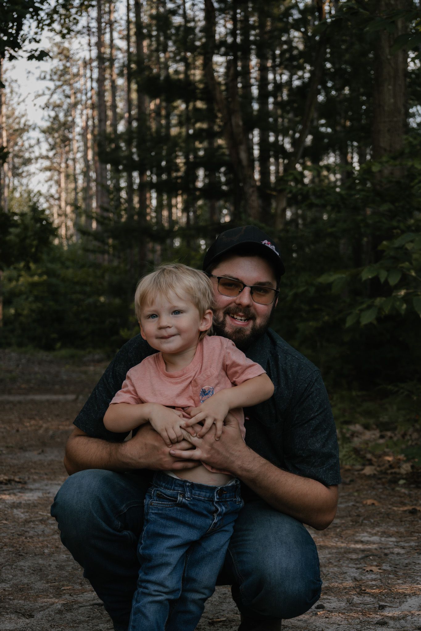 A man crouching in a forest hugging a young blonde child, both smiling outdoors among tall trees.