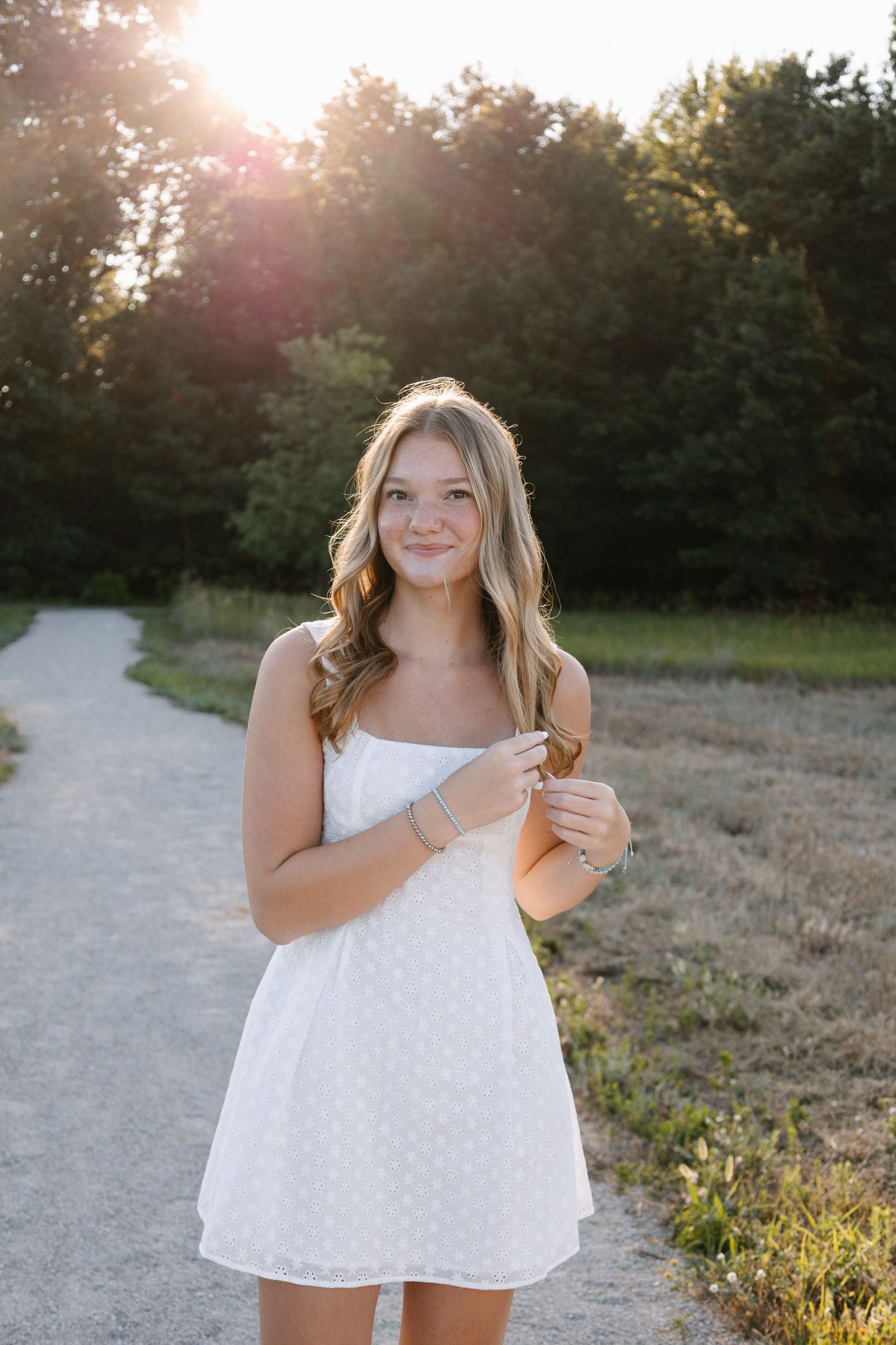 Young woman in white sundress smiling on a path at golden hour with trees in background.