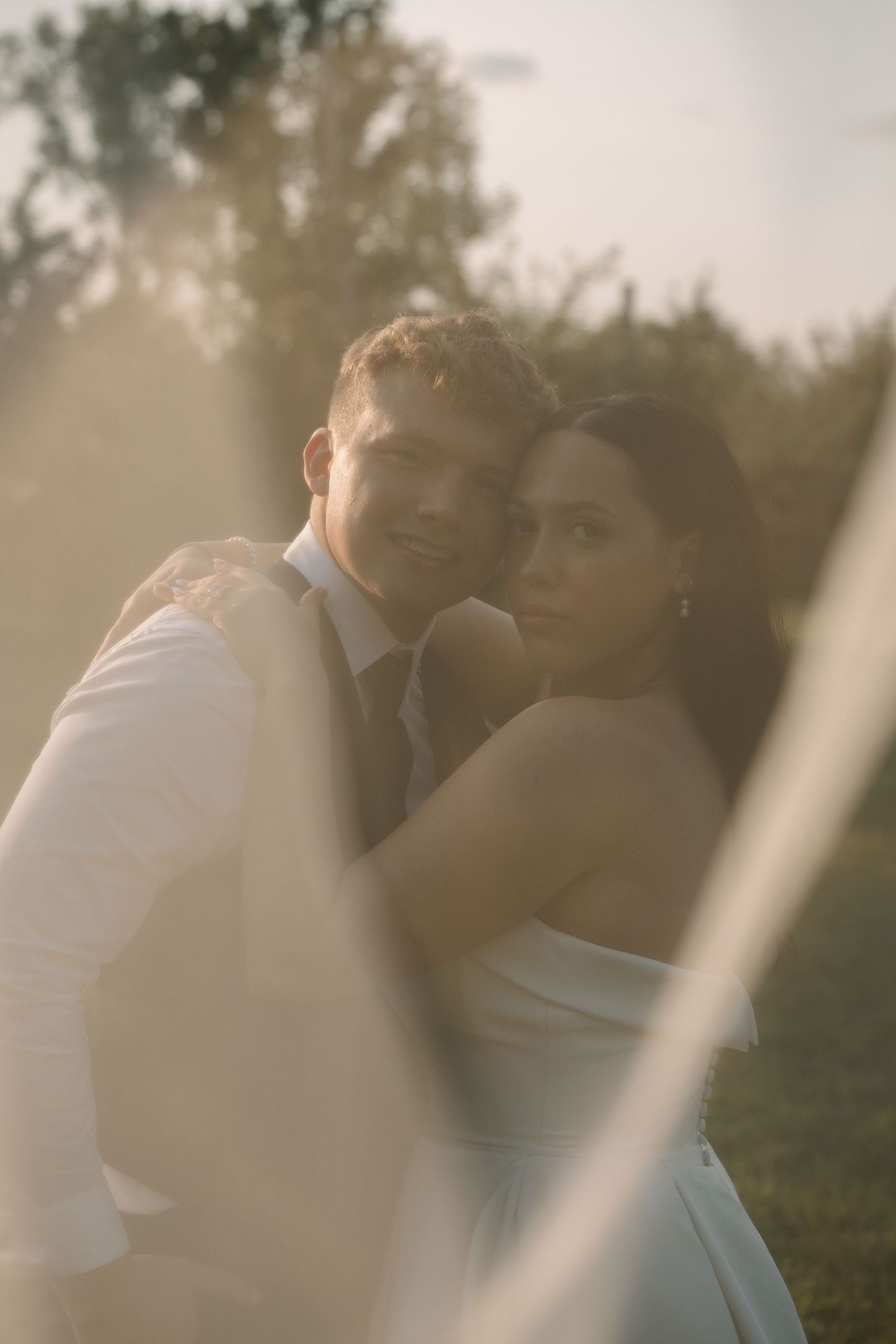 Bride and groom share a romantic golden hour portrait through a soft veil in lush greenery.