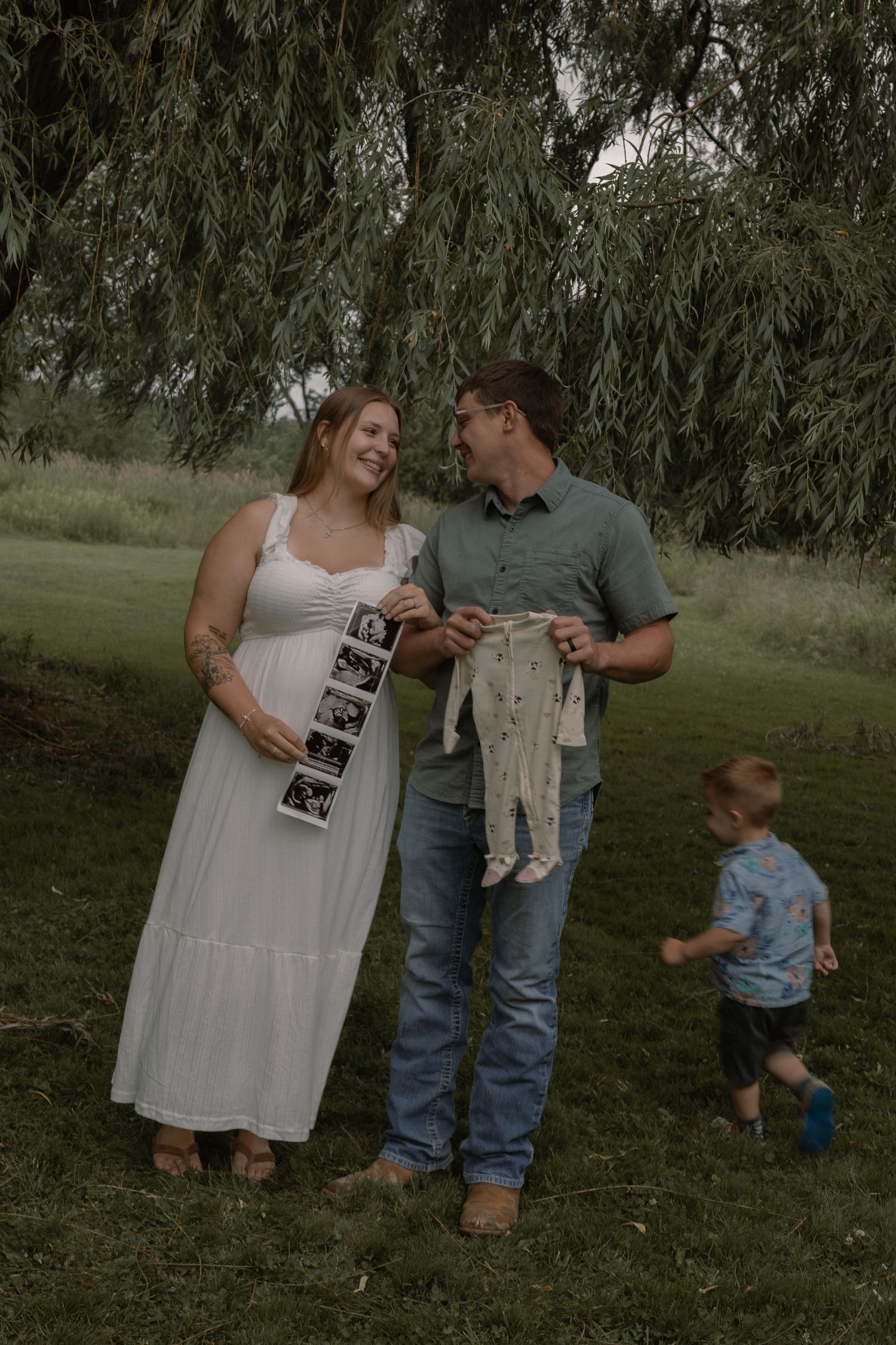 Expecting couple holding ultrasound photos and baby onesie while toddler plays nearby under willow tree.
