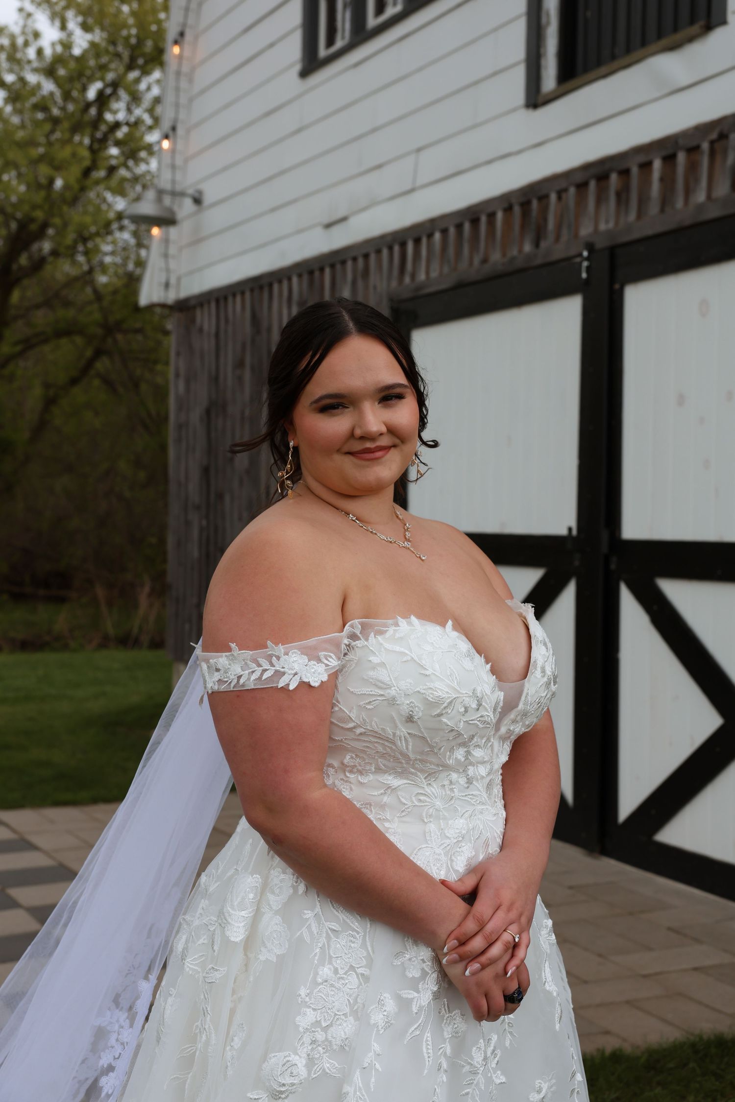 Smiling bride in strapless lace off-shoulder wedding gown with veil standing outside rustic white barn.