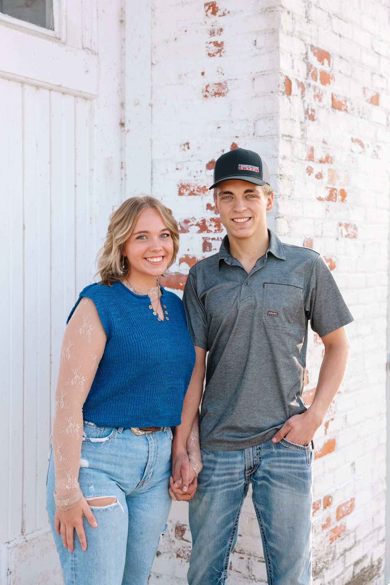 A smiling couple holds hands while standing together against a rustic white brick wall outdoors.